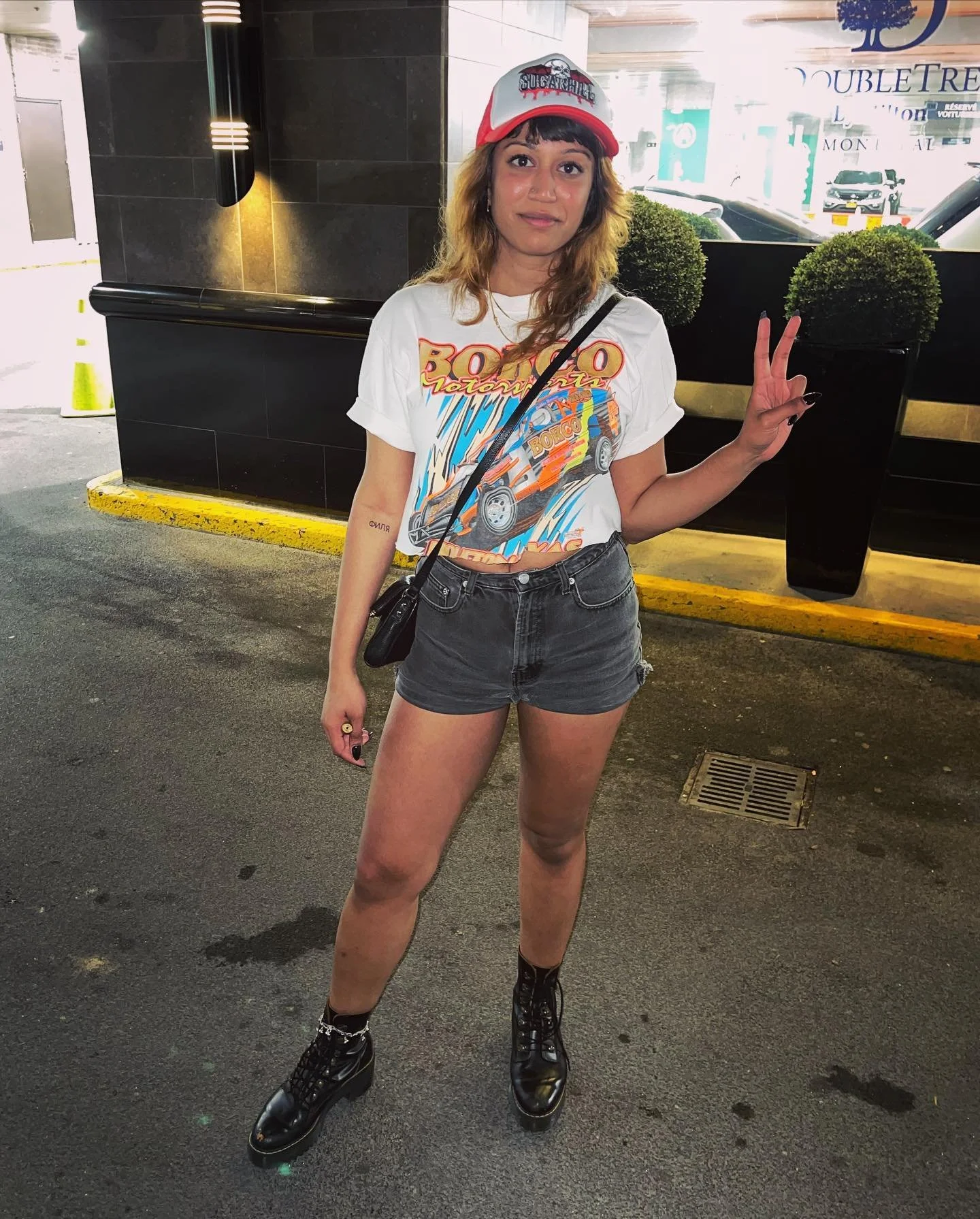 Person wearing a graphic tee, denim shorts, and boots, posing with a peace sign in front of a DoubleTree hotel entrance.