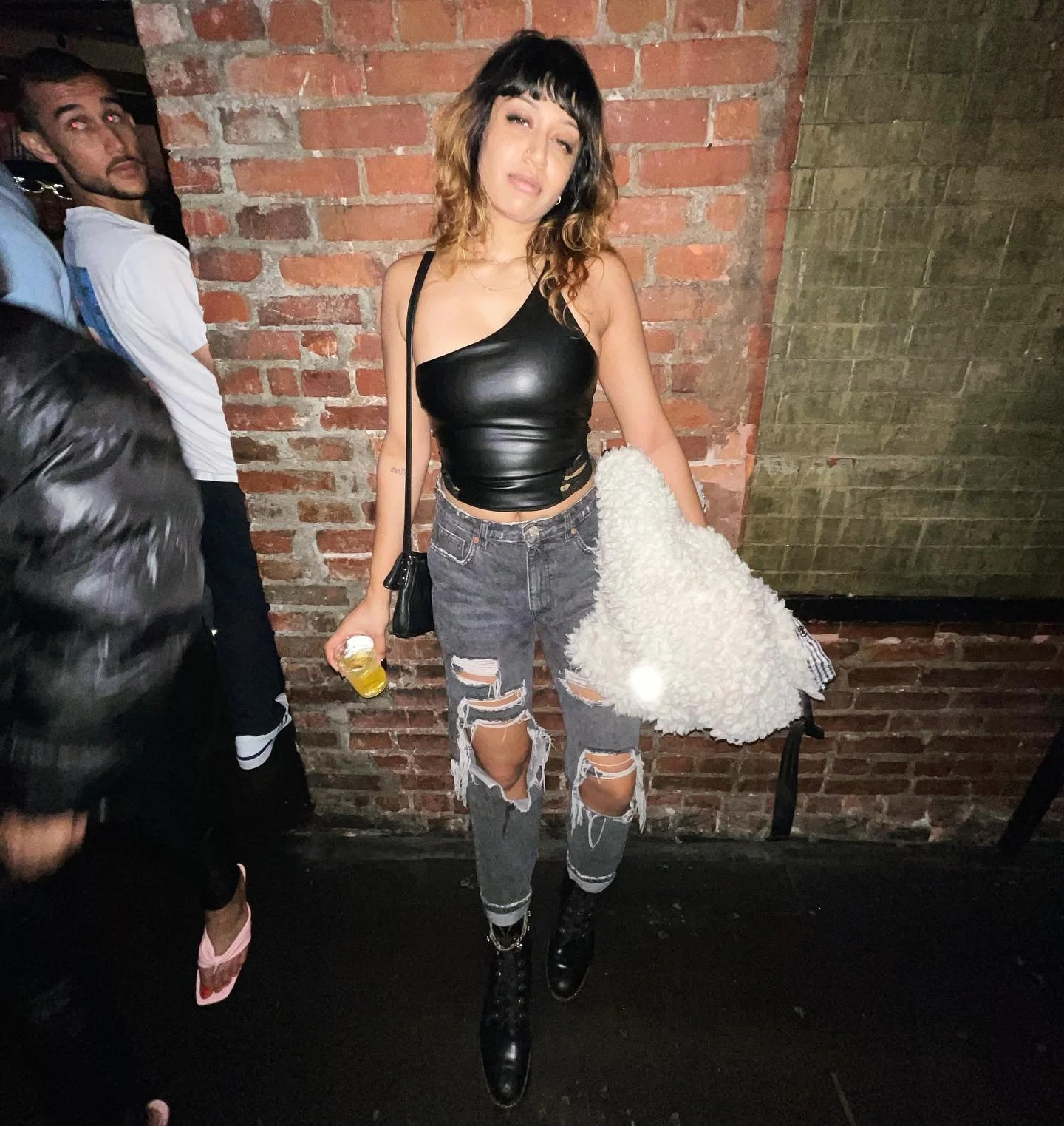 Woman in black leather top, ripped jeans holding a drink in a bar with brick walls, casual style