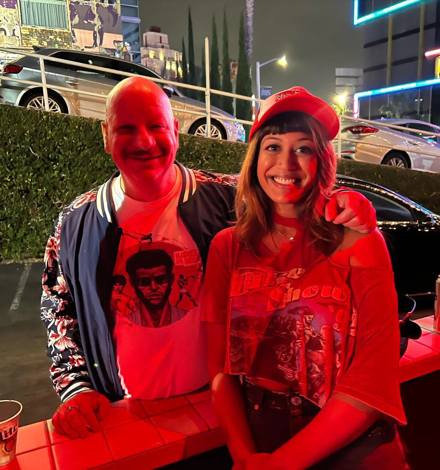 Two people smiling at night, with a parking area in the background, wearing casual graphic tees and a red baseball cap.