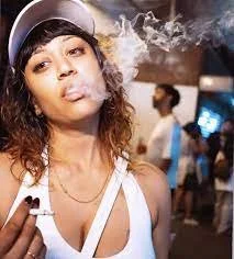 Person smoking in a casual outdoor setting, wearing a white tank top and cap, with visible smoke.