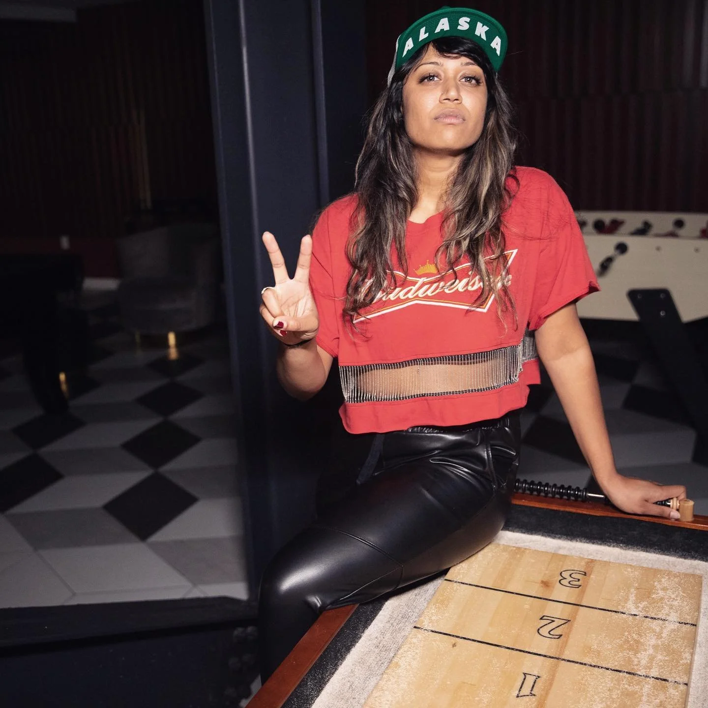 Person wearing a red shirt and an 'Alaska' cap making a peace sign, sitting on a shuffleboard table.