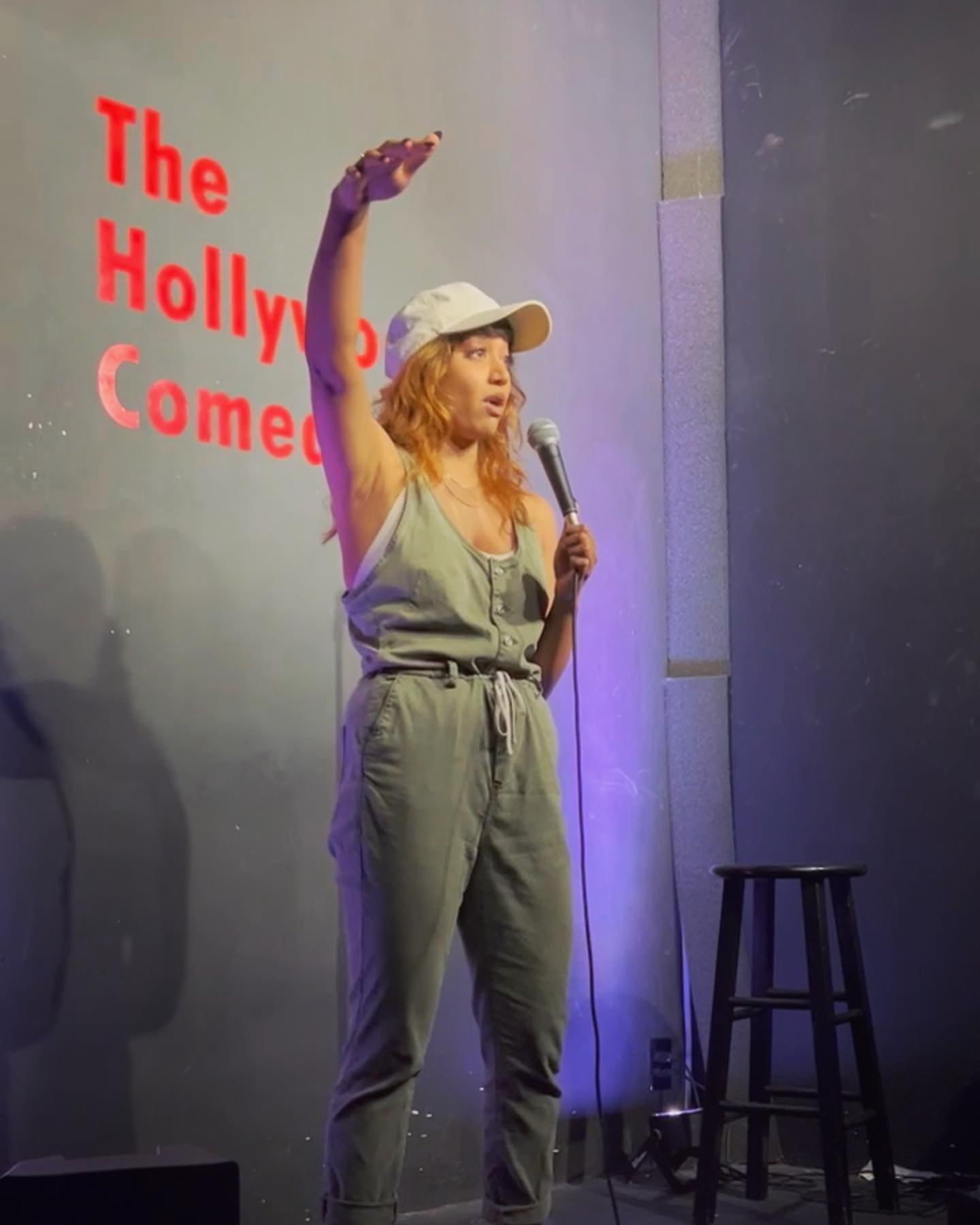 Person wearing a cap and casual clothing performing stand-up comedy on stage with a microphone, next to a stool. "The Hollywood Comedy" sign in the background.