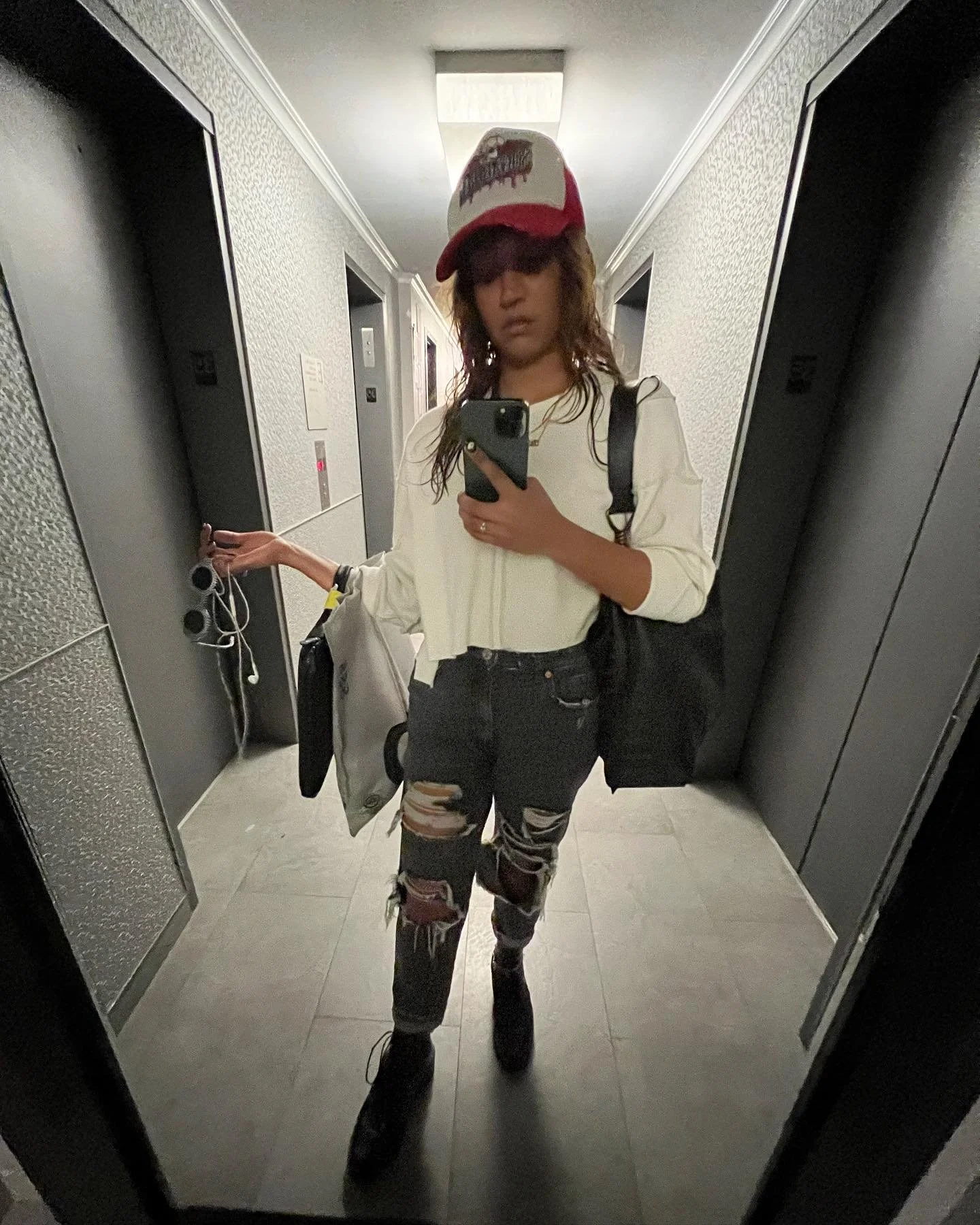 A woman taking a mirror selfie in a hallway, wearing a red and white cap, white sweatshirt, distressed jeans, and black boots, holding a phone and a white bag.