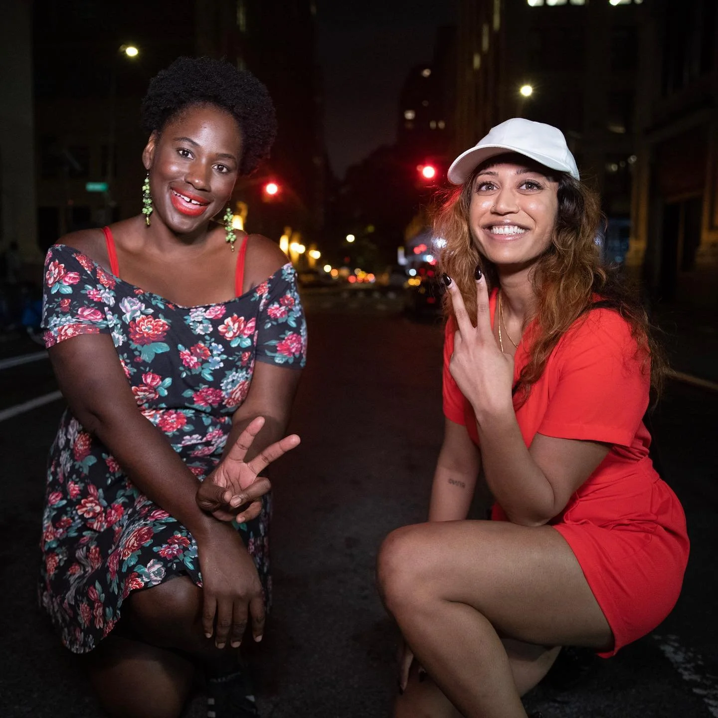 Two women kneeling and smiling at night on a city street. The woman on the left is wearing a floral dress, and the woman on the right is wearing a red outfit and a white cap. Both are gesturing peace signs with their hands.