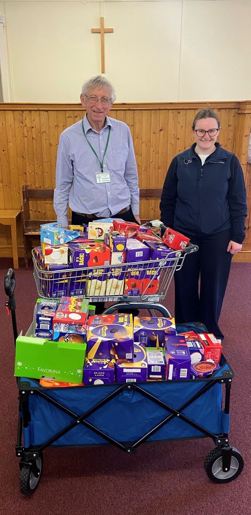 Two people, a man and a woman, standing behind a shopping cart filled with grocery items, in a room with wood paneling and a cross on the wall.