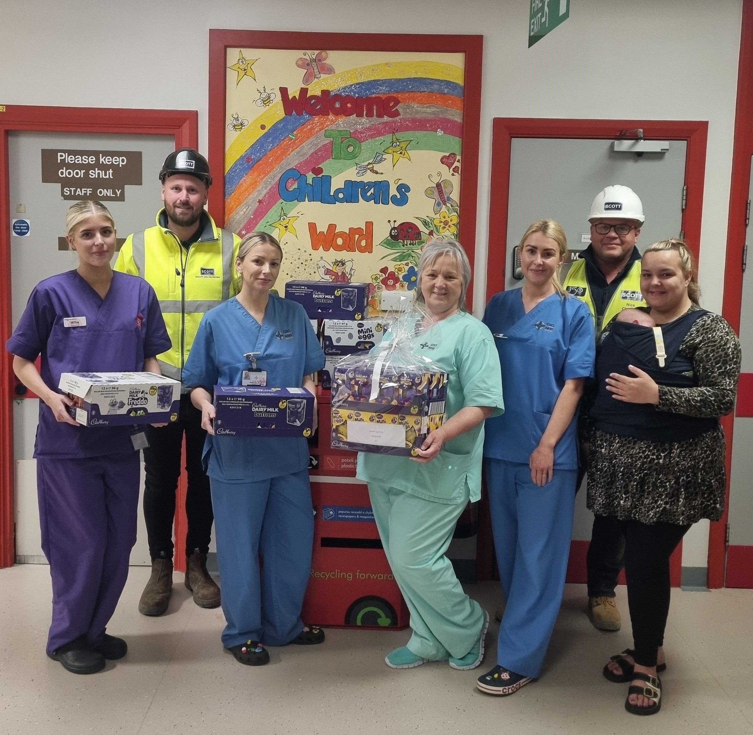 Group of healthcare workers and adults standing with children in front of a colorful sign that reads 'Welcome to Children's Ward,' holding gift boxes. The setting appears to be inside a hospital or healthcare facility.
