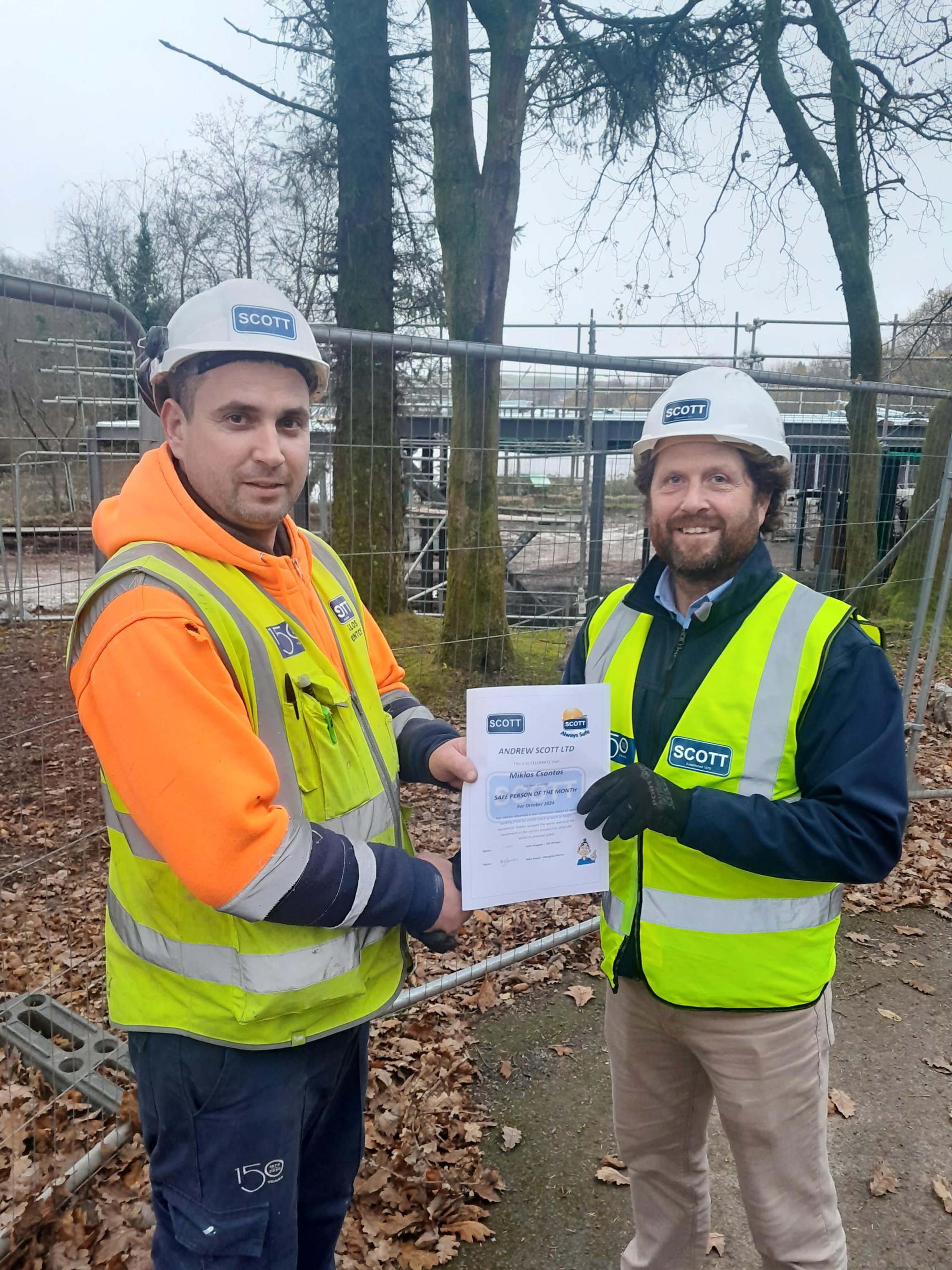 Two construction workers wearing high visibility vests and hard hats, standing outdoors with leaf-covered ground, holding a certificate of recognition.