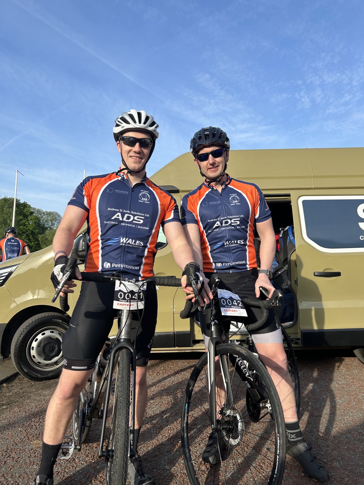 Two male cyclists in matching team jerseys standing with their bikes in front of a yellow vehicle during a race event, smiling at the camera.