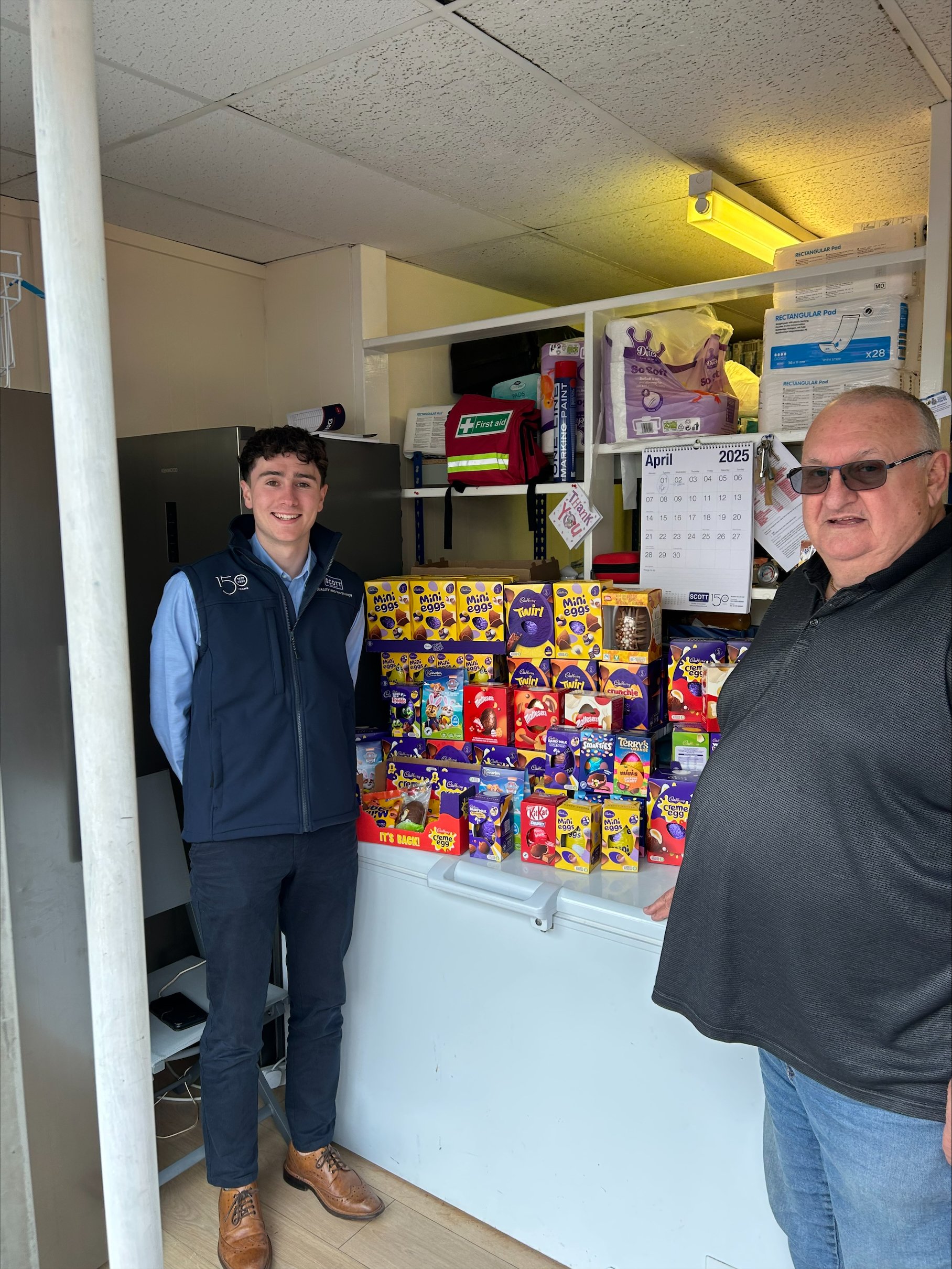 Two men standing in a small store or storage room, with shelves filled with various items including Holiday-themed candy boxes and online shopping boxes, smiling at the camera.