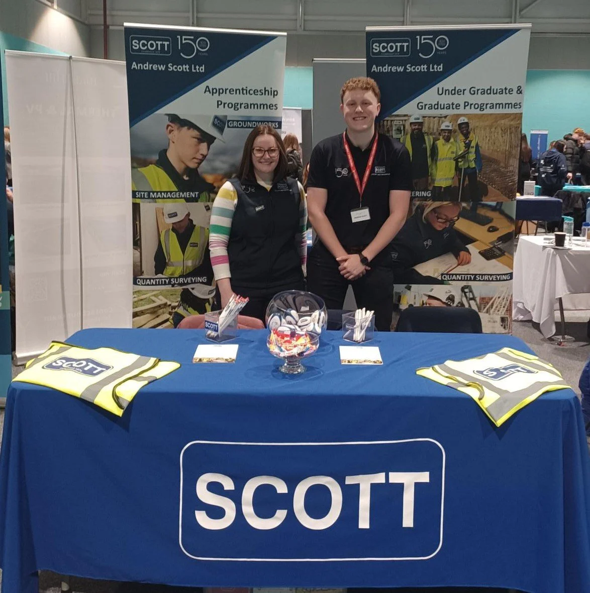 Two people standing behind a table at a Scott Ltd booth displaying promotional material and branded items, with banners advertising apprenticeship and graduate programs in construction and surveying.
