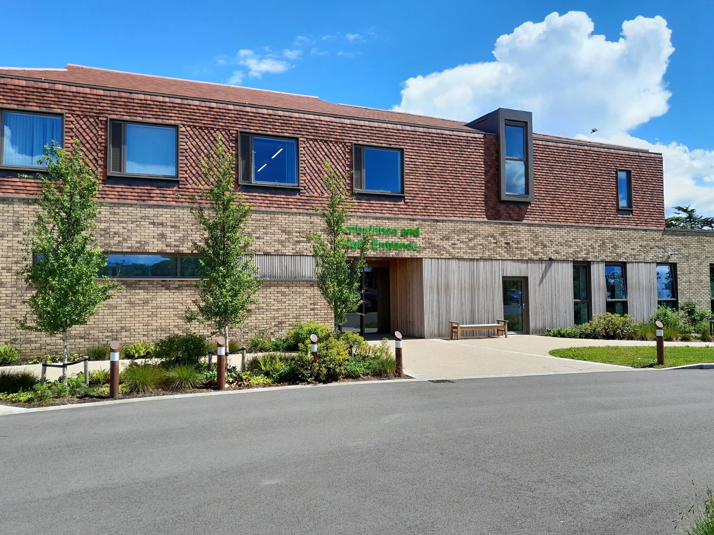 Modern two-story building with brick and wood facade, large windows, and surrounded by landscaping, under a blue sky with clouds.