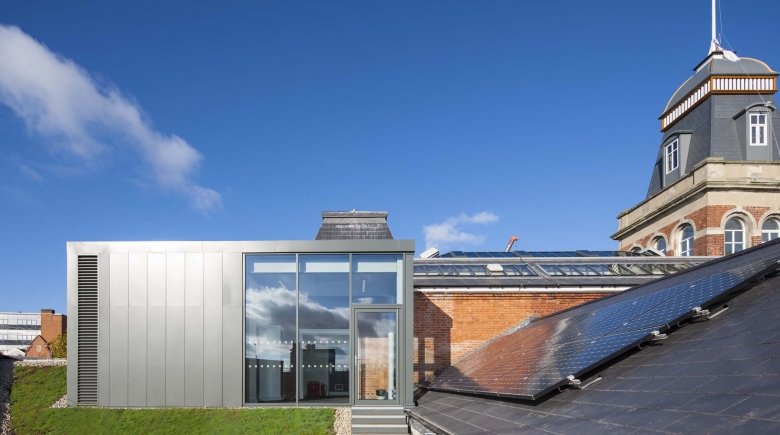 Modern building with large glass windows and metal exterior, adjacent to an older brick building with a tower and solar panels on the roof, under a partly cloudy blue sky.