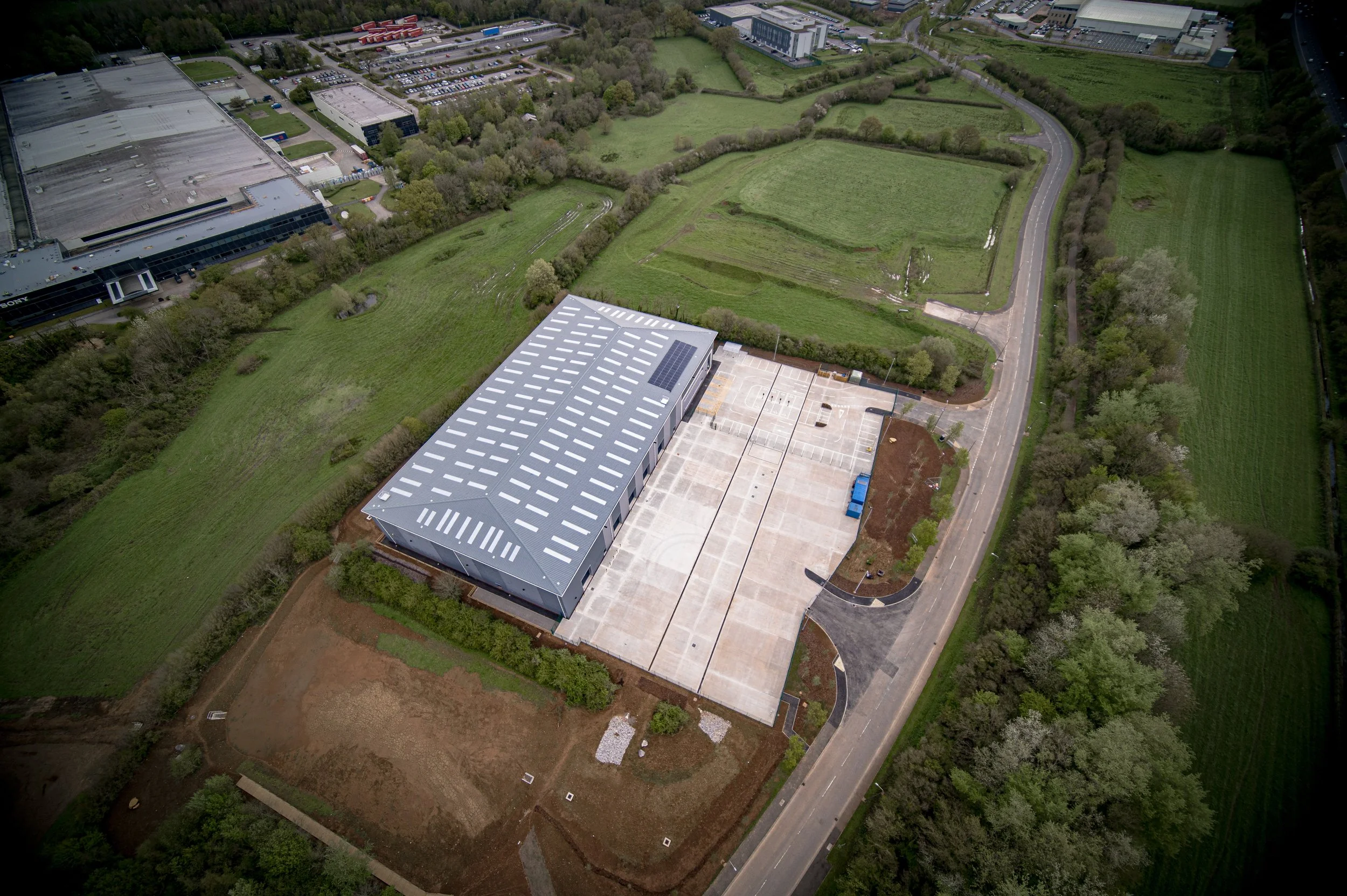 An aerial view of a large industrial building with a solar panel on its roof, surrounded by parking lots and green fields, with a road running along the side.