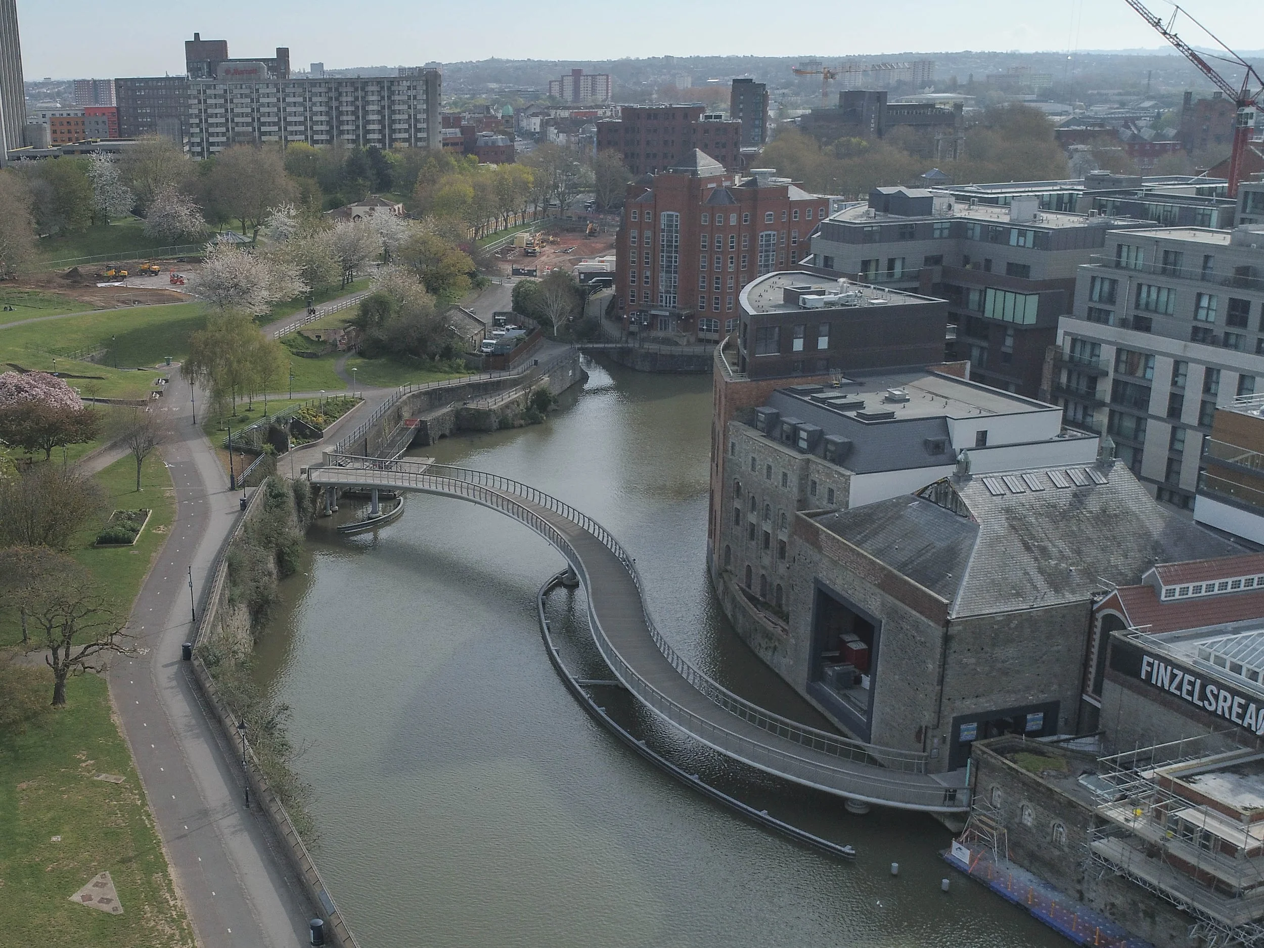 A cityscape with a river and a curved pedestrian bridge, surrounded by trees, green spaces, and modern buildings under a cloudy sky.