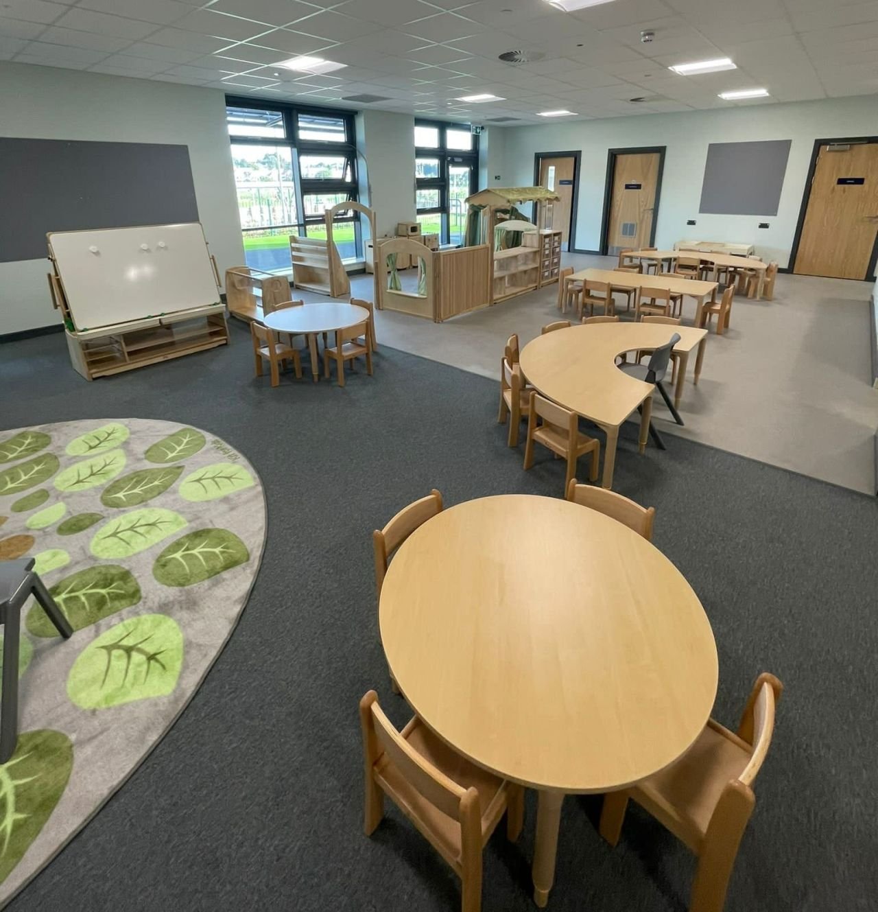 Preschool classroom with wooden tables and chairs, educational play areas, whiteboard, and circular rug with leaf pattern.