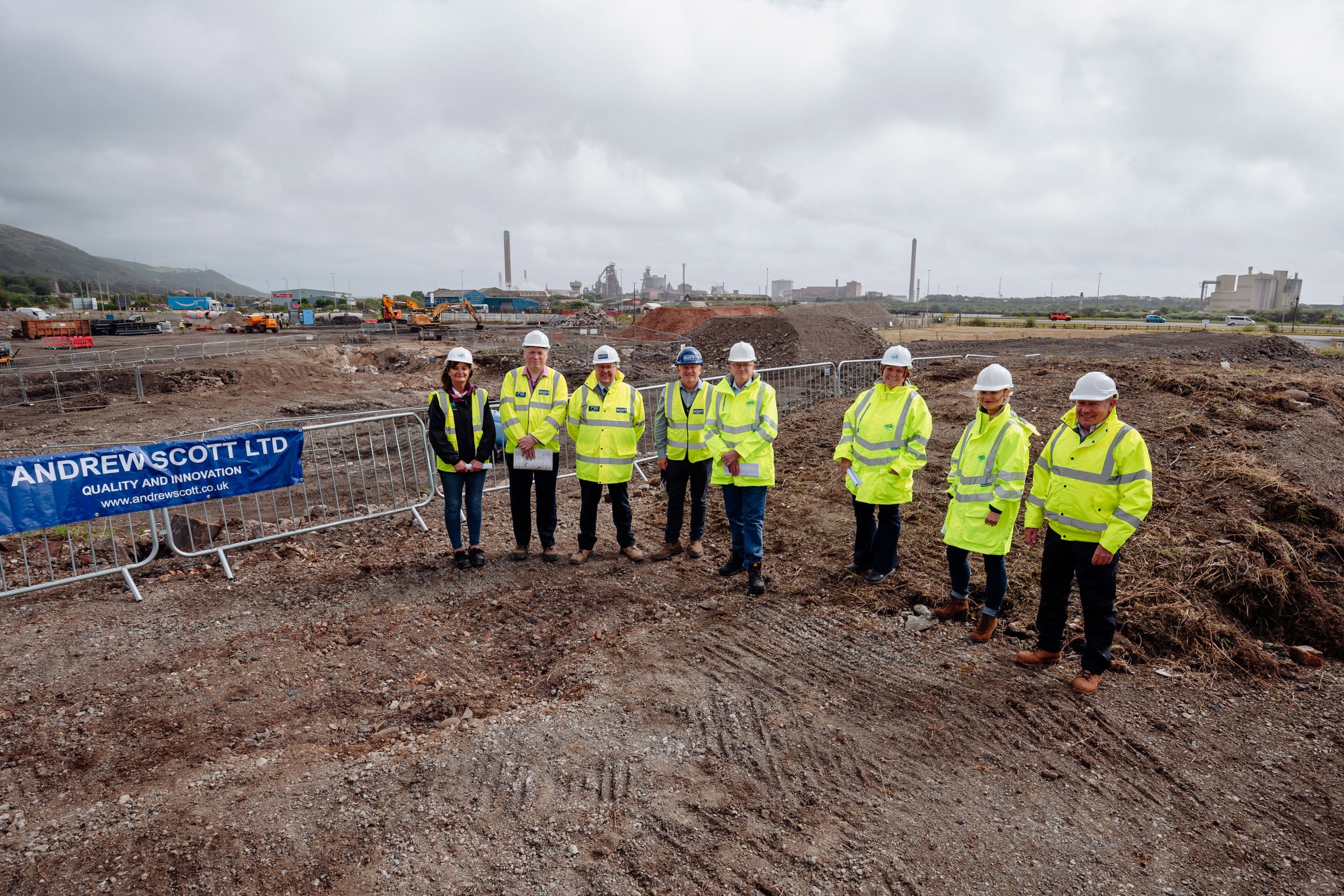 Group of eight people wearing yellow high-visibility jackets and white safety helmets standing on a construction site with excavated dirt, construction equipment, and industrial buildings in the background.