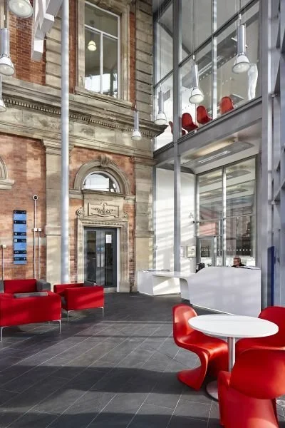 Modern lobby with glass walls, red chairs, and a reception desk inside a building with an exposed brick exterior.