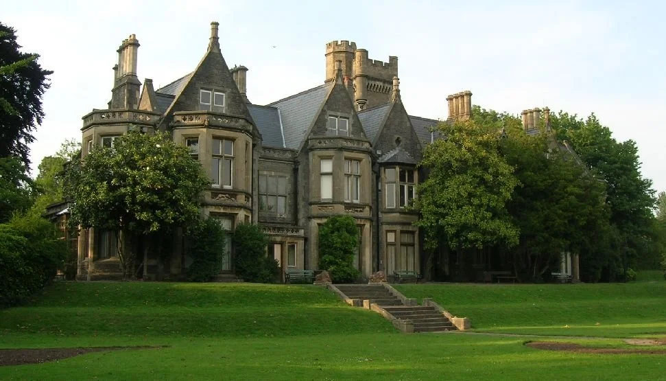 A large, historic mansion with multiple turrets and pointed roofs, surrounded by trees and a well-maintained lawn.