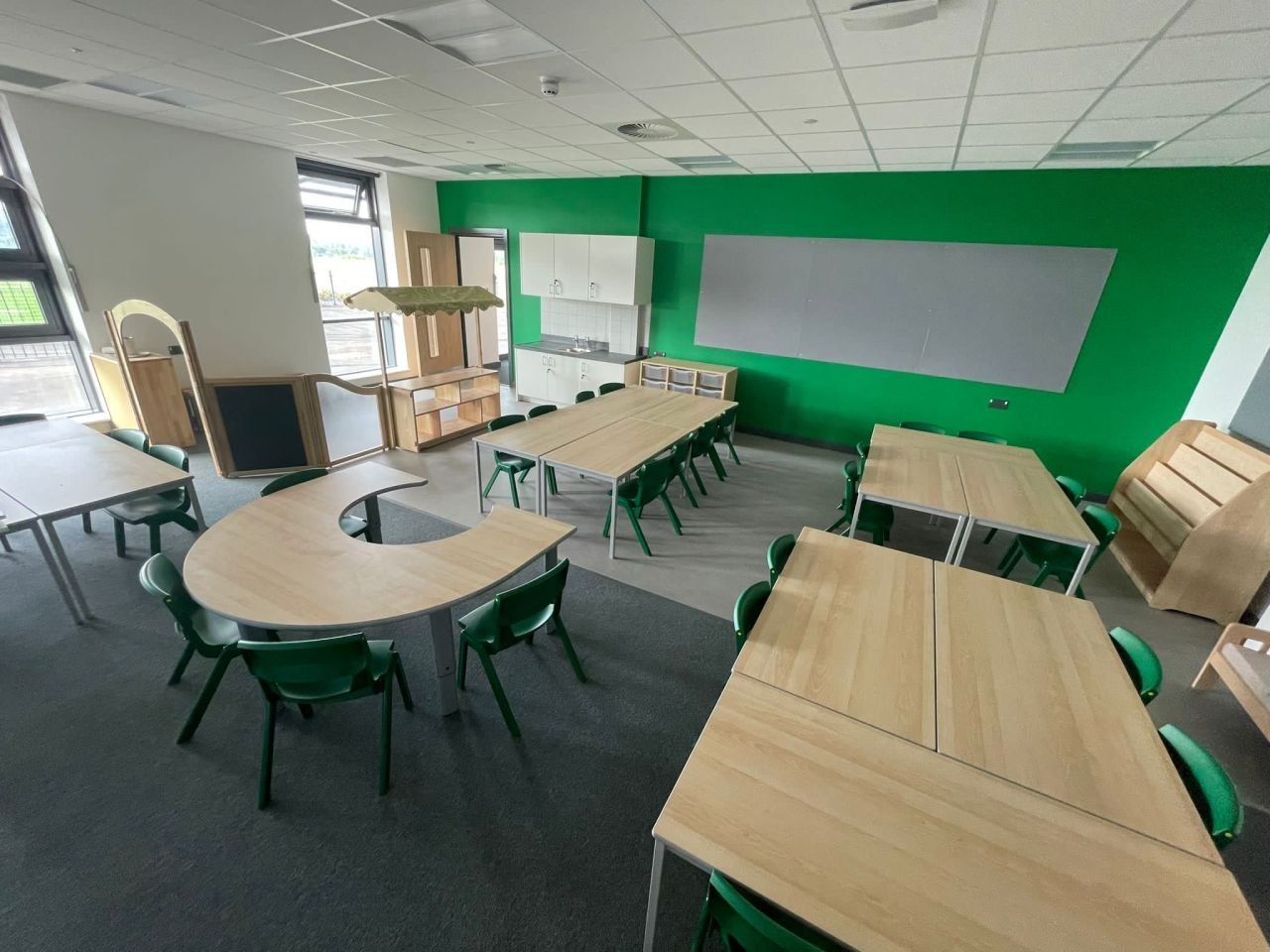 Empty classroom with wooden tables and green chairs, green wall, cabinets, and large windows.