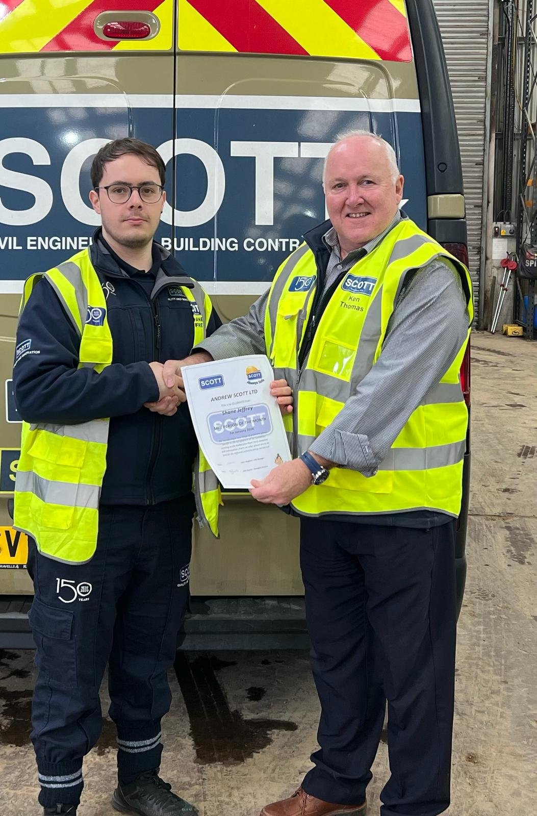 Two men in high visibility vests shaking hands, one holding a certificate, standing in front of a work van marked with 'SCOTT' and related construction branding.