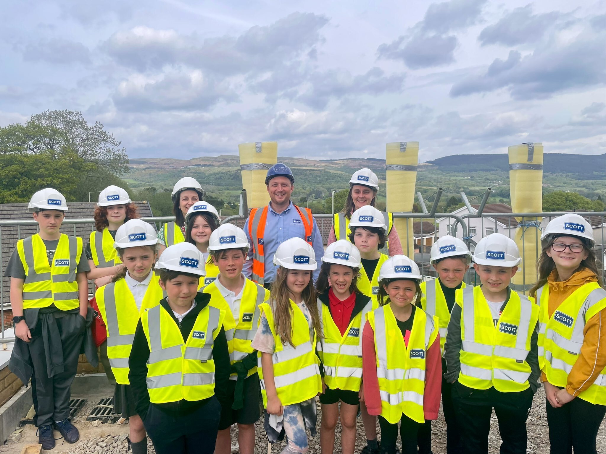 Group of children and adults wearing safety helmets and high-visibility vests on a construction site, with a scenic landscape in the background.