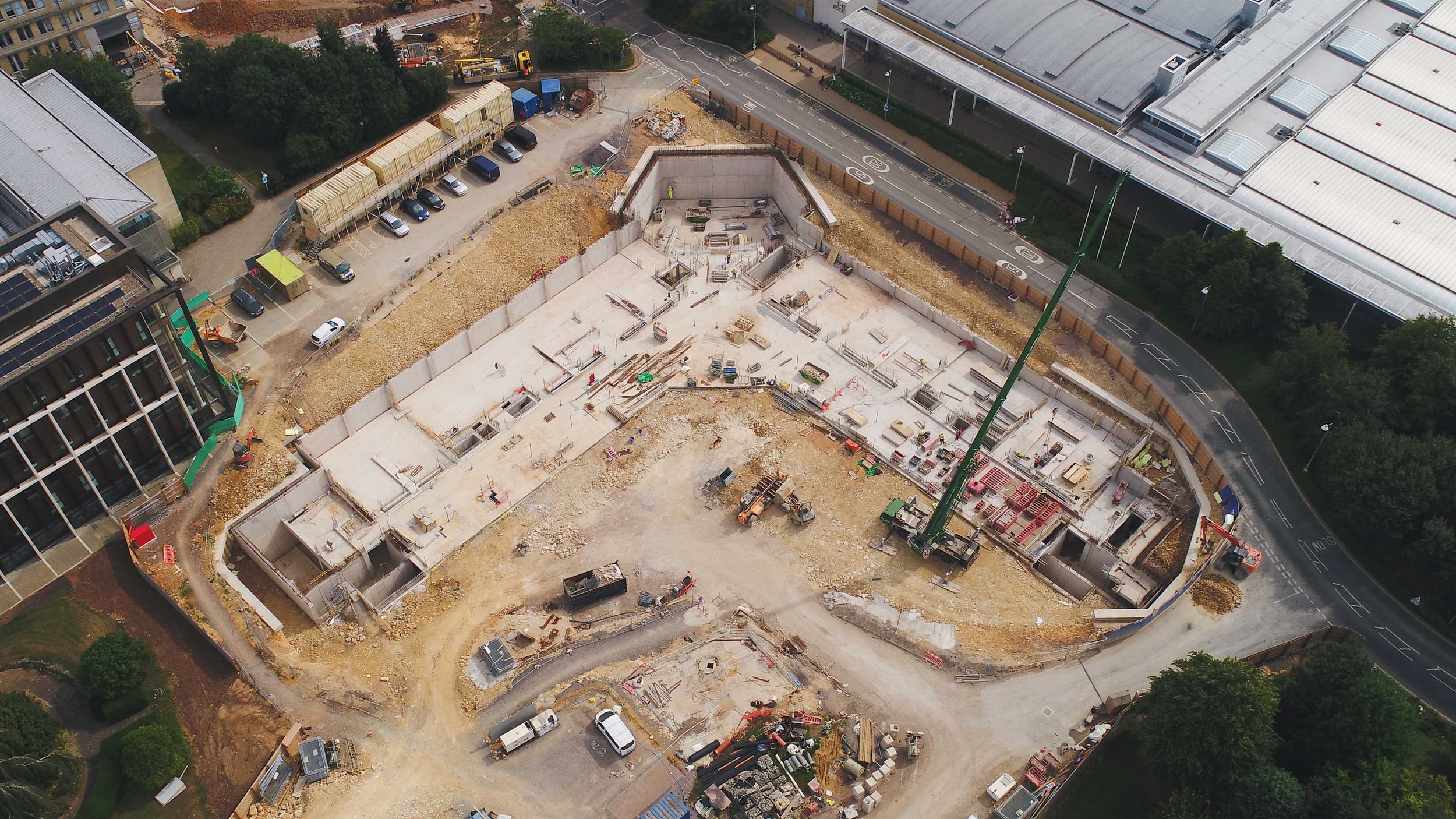 Aerial view of a construction site with partially built concrete structures, construction equipment, and vehicles, surrounded by roads, parking lots, and nearby buildings.