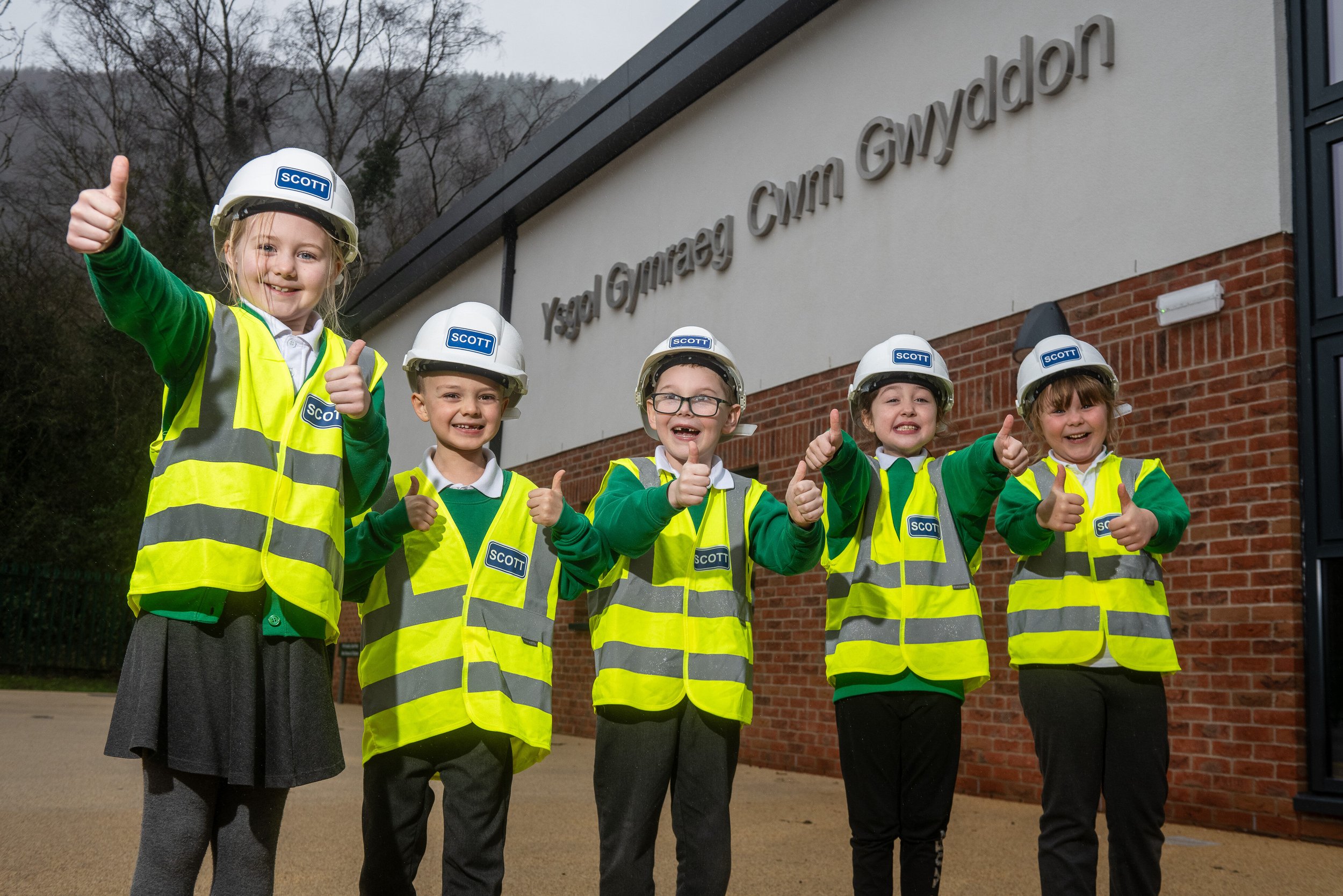 Five children wearing yellow safety vests and white construction helmets giving thumbs-up in front of a building with a sign.