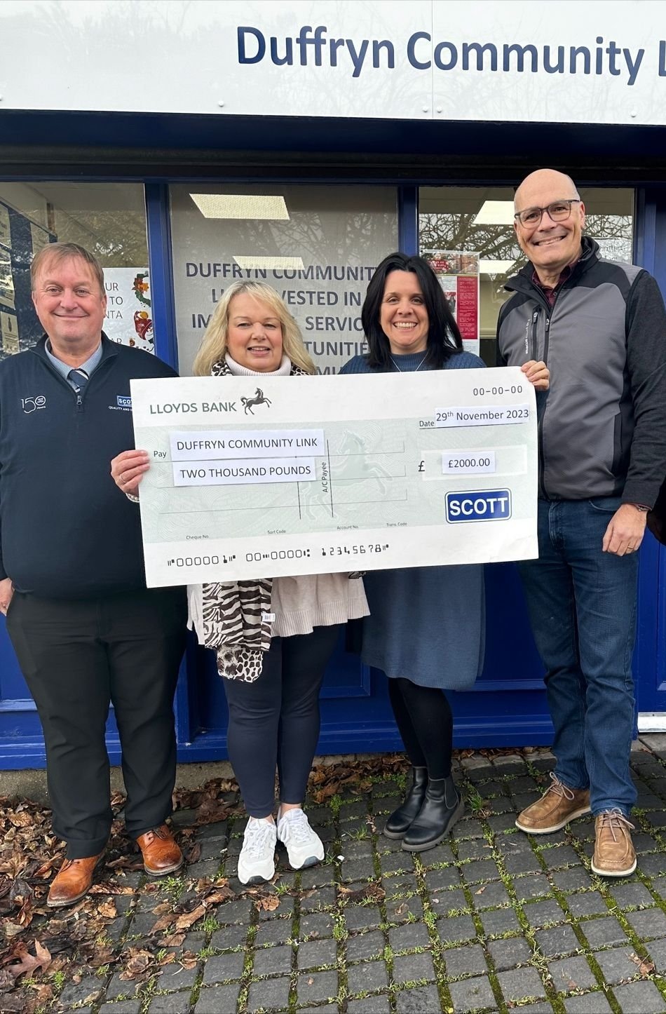 Four individuals standing outside Duffryn Community Link, holding an oversized check from Lloyds Bank for 2000 pounds, dated November 29, 2023, celebrating a donation. The individuals are smiling, dressed in casual to semi-formal attire, and standing