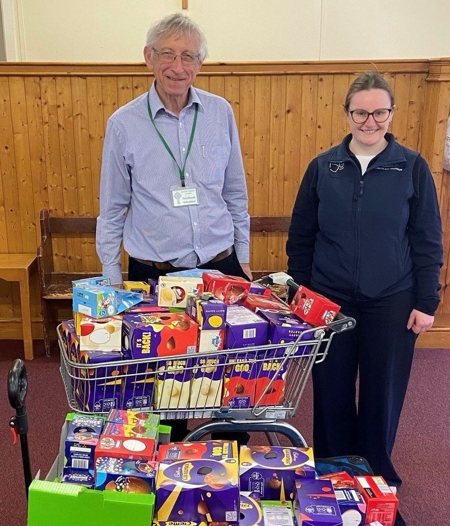 Two people, an older man and a woman, stand behind a shopping cart filled with boxes of chocolates, in a wood-paneled room, smiling at the camera.