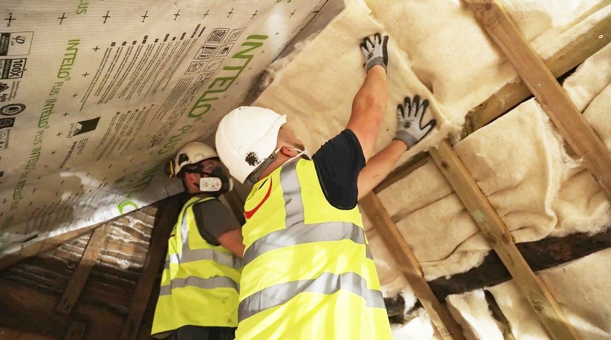 Workers installing or inspecting insulation in an attic or ceiling space, wearing safety helmets and high-visibility vests.