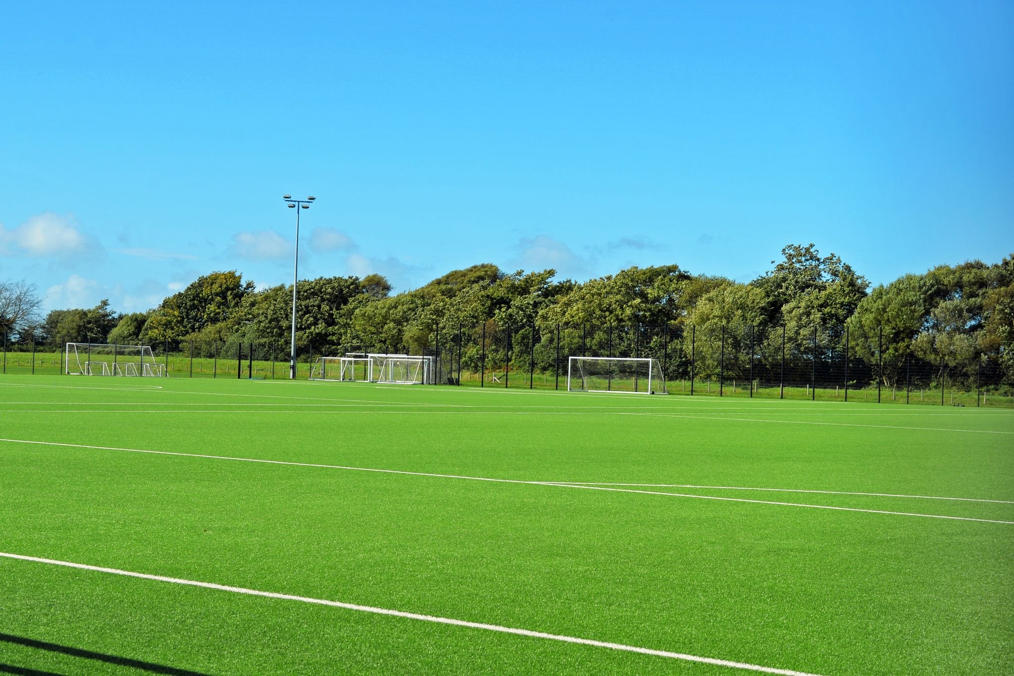 Outdoor soccer field with goalposts, green artificial turf, and surrounding trees under a clear blue sky.