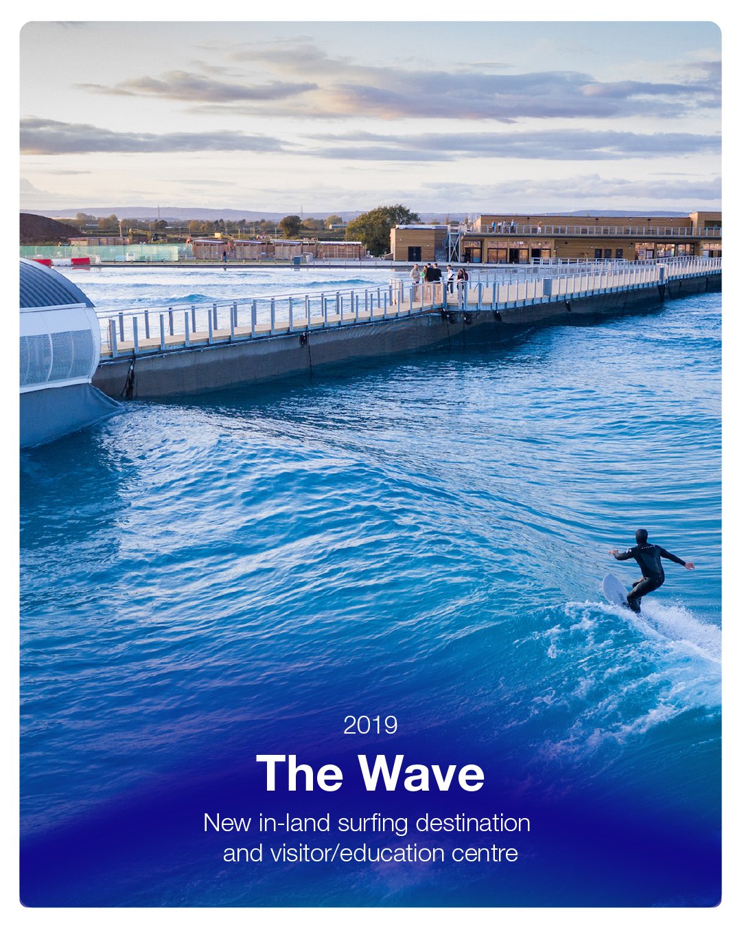 Photo of an in-land surfing attraction called 'The Wave' with a person surfing on artificial water waves, a walkway with visitors, and a building in the background, under a cloudy sky.