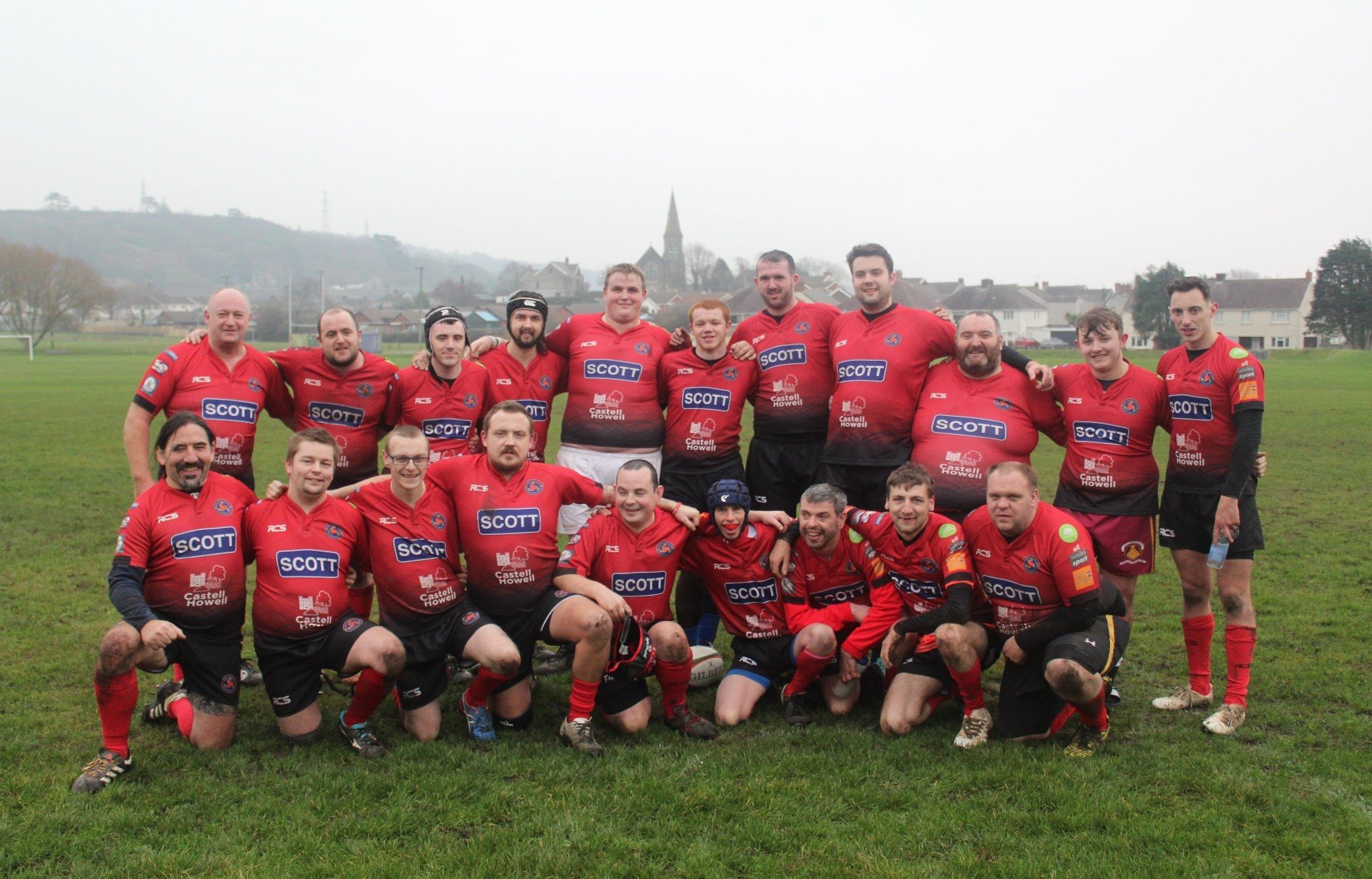 Group of rugby players wearing red jerseys and black shorts on a grass field, posing for a team photo. Some players are kneeling in front, while others are standing in the back, with arms around each other's shoulders, on an overcast day.