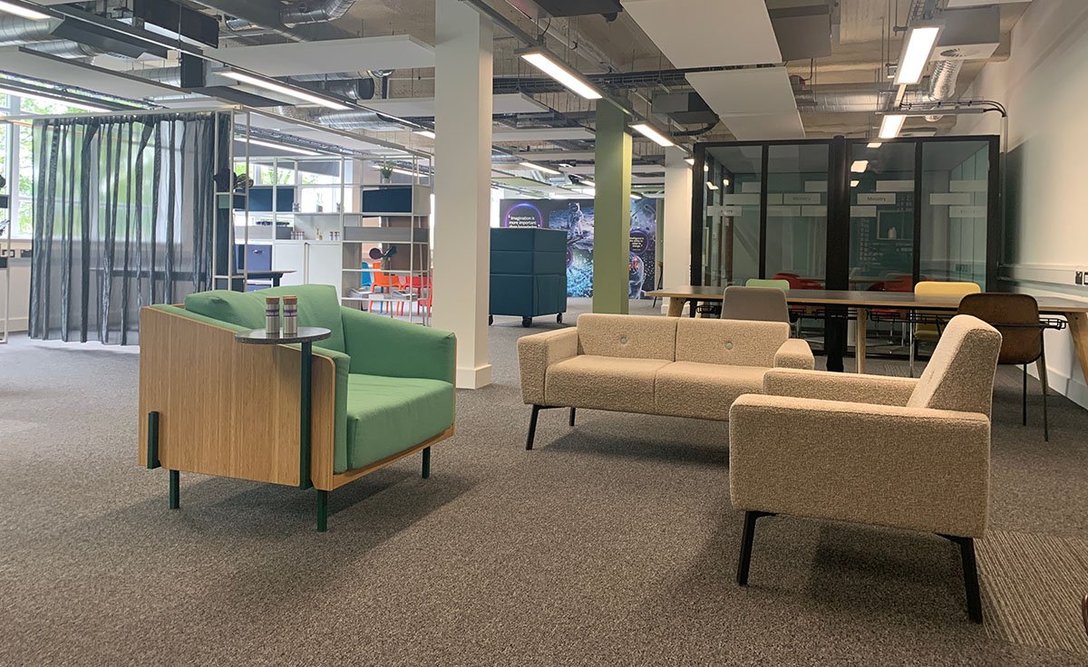 Modern office lounge area with green and beige sofas, a table with colorful chairs, and an industrial ceiling design.