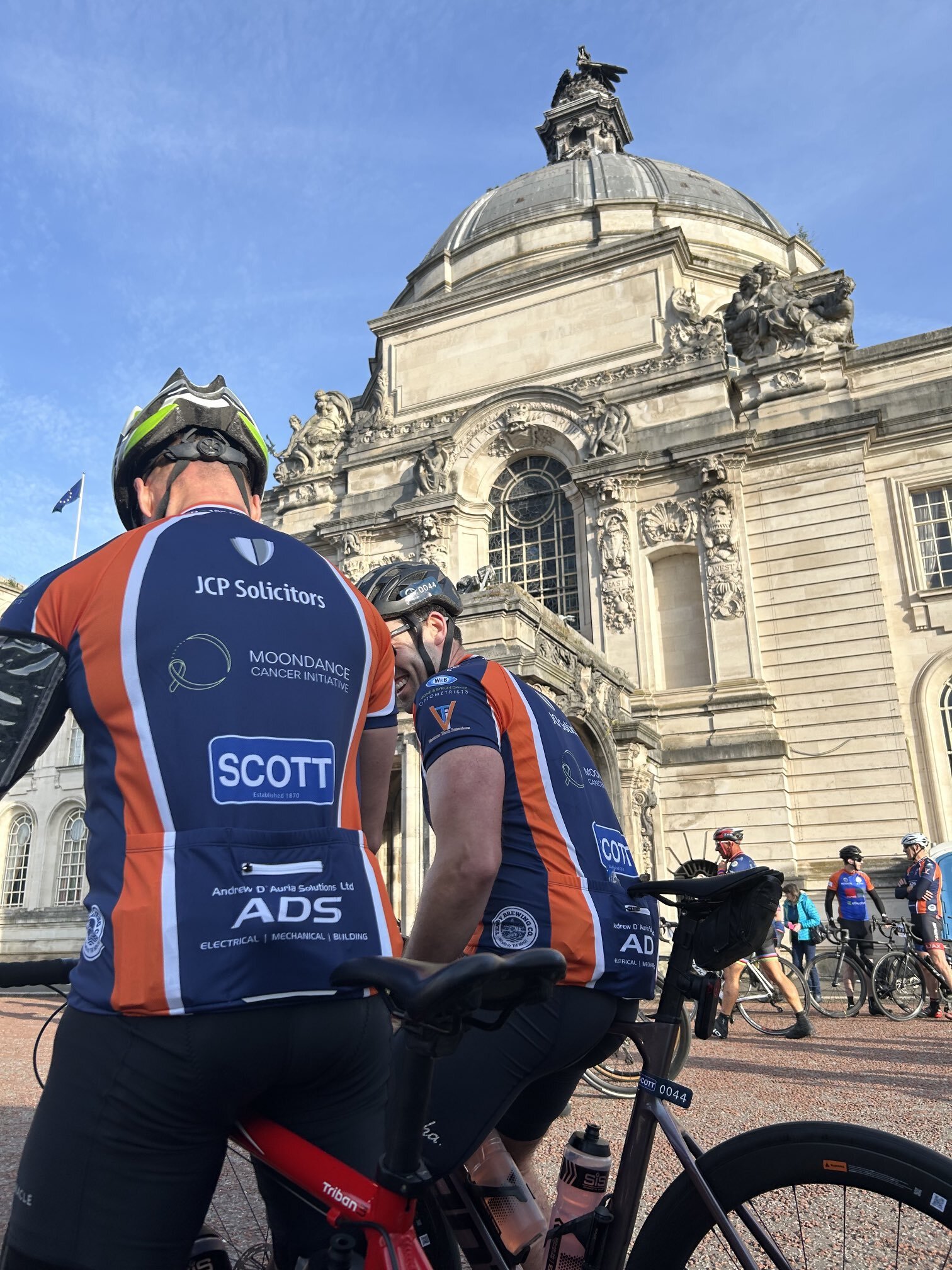 Cyclists wearing matching jerseys gather in front of an ornate historic building with a domed roof and detailed sculptures.