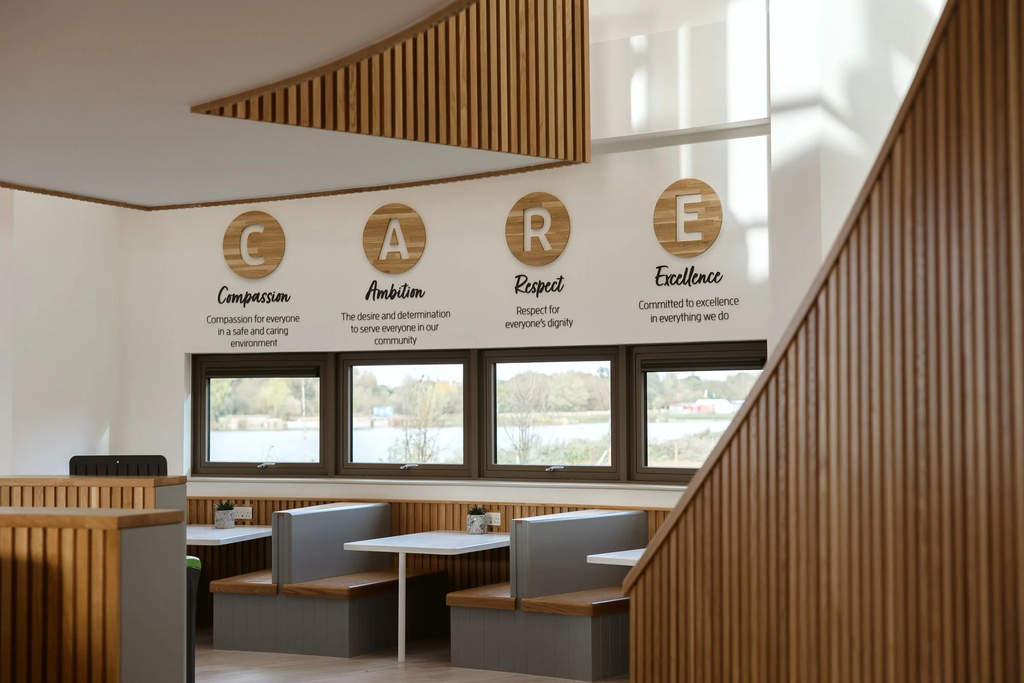 Interior of a modern room with large windows, wooden decor, and a wall display spelling CARE with each letter representing a value: Compassion, Ambition, Respect, Excellence.