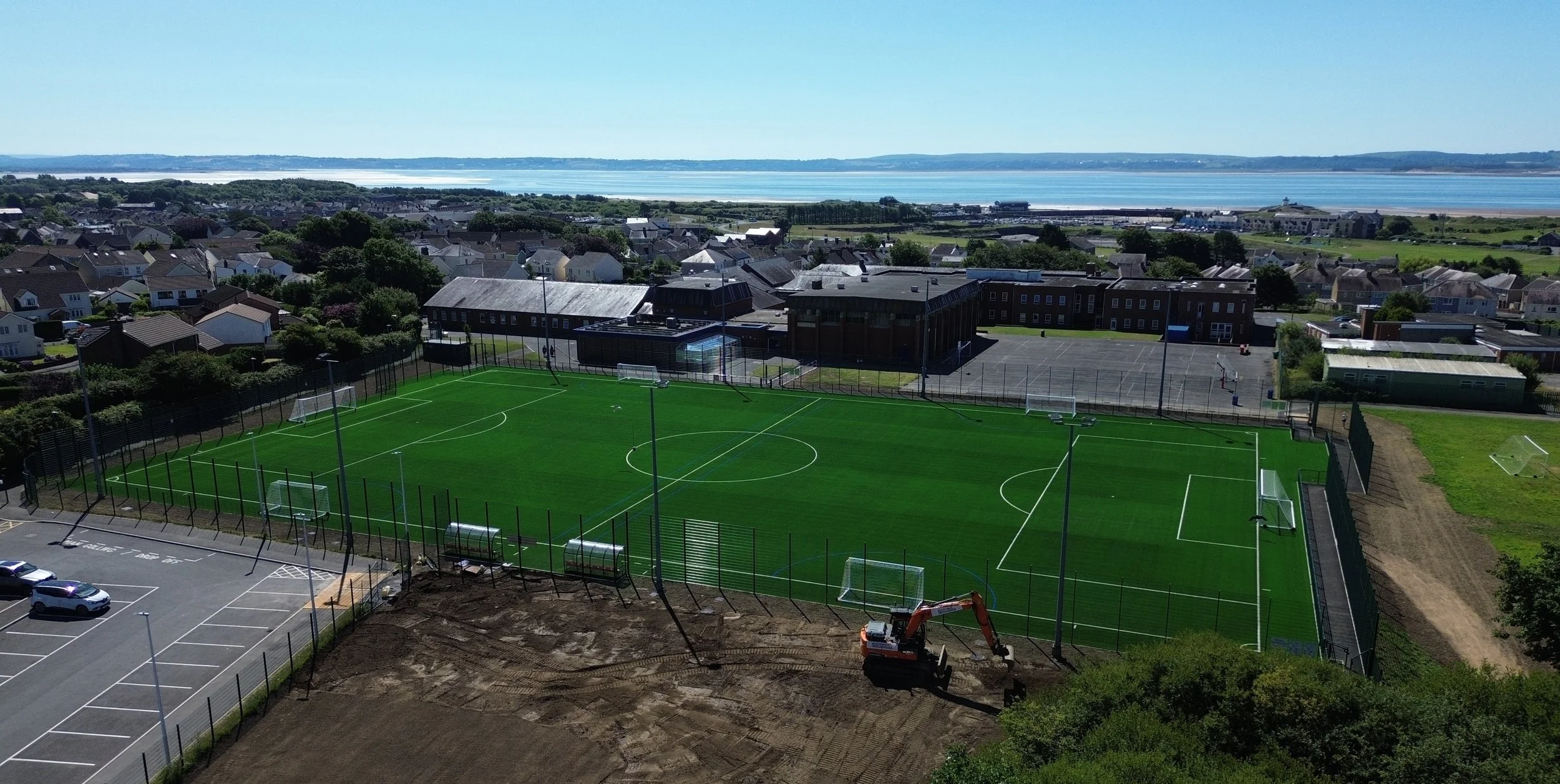 A soccer field under construction with artificial turf, surrounded by a black fence, with construction equipment nearby and parking lot on the left, in a coastal town with residential houses and a body of water in the background.