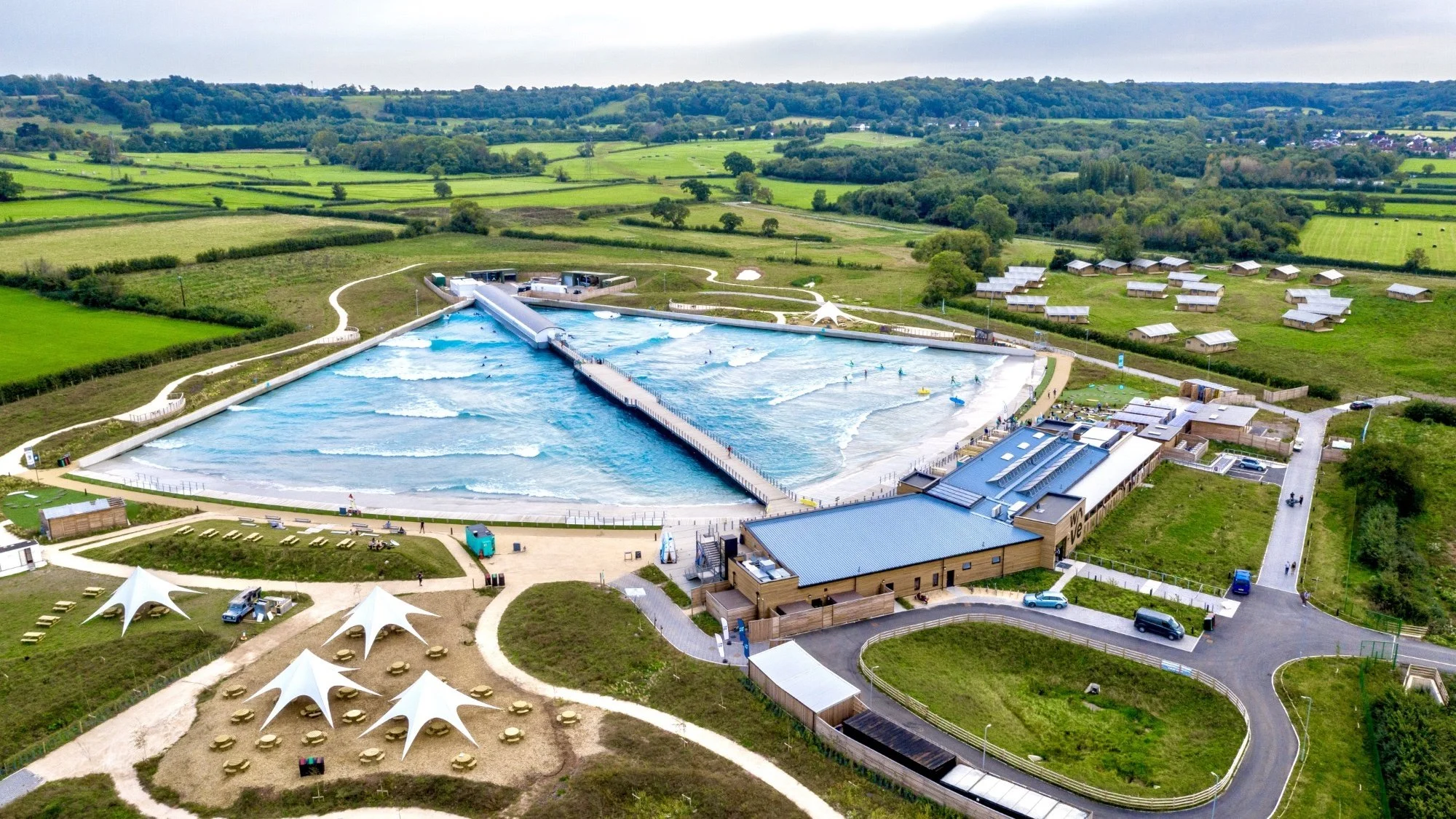 Aerial view of an artificial wave pool surrounded by green fields and countryside, featuring tents and buildings nearby.