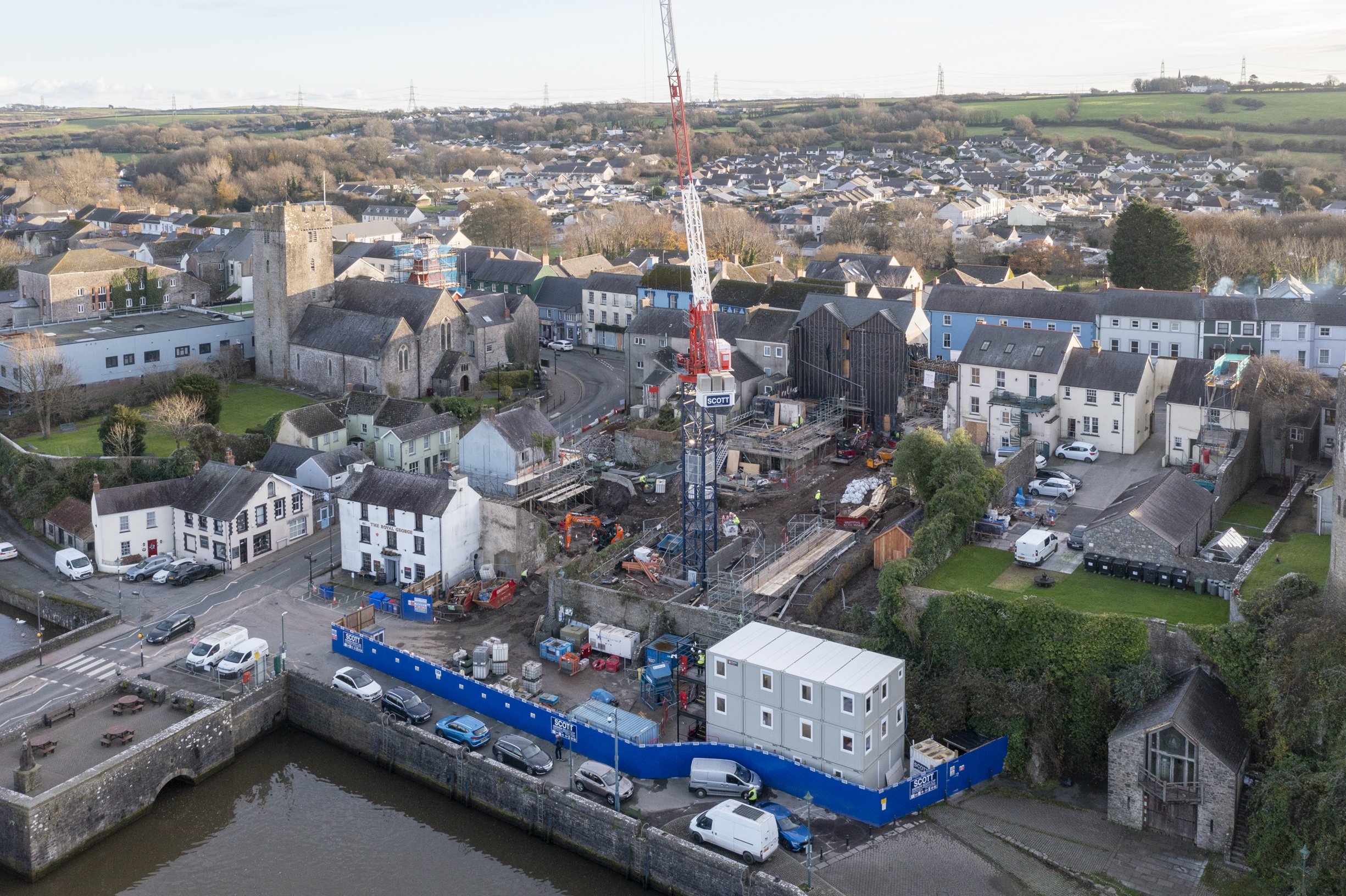 Aerial view of a construction site in a town with a crane, surrounded by historic buildings, a church, residential houses, and a riverfront.