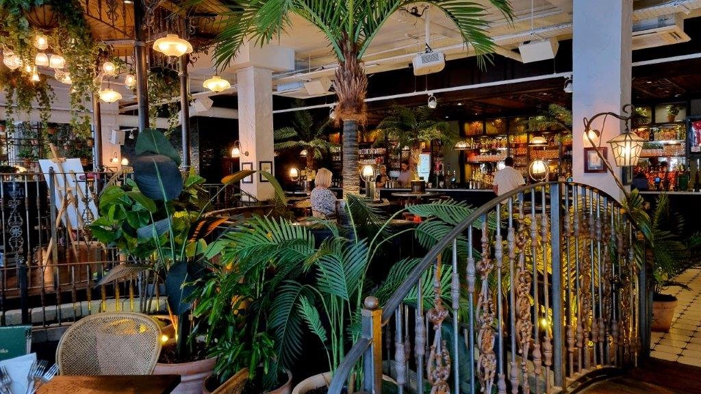 Interior of a stylish bar with lush greenery, decorative lights, and a curved iron railing. Several people are seated at the bar counter, and the space is adorned with hanging plants and warm lighting.