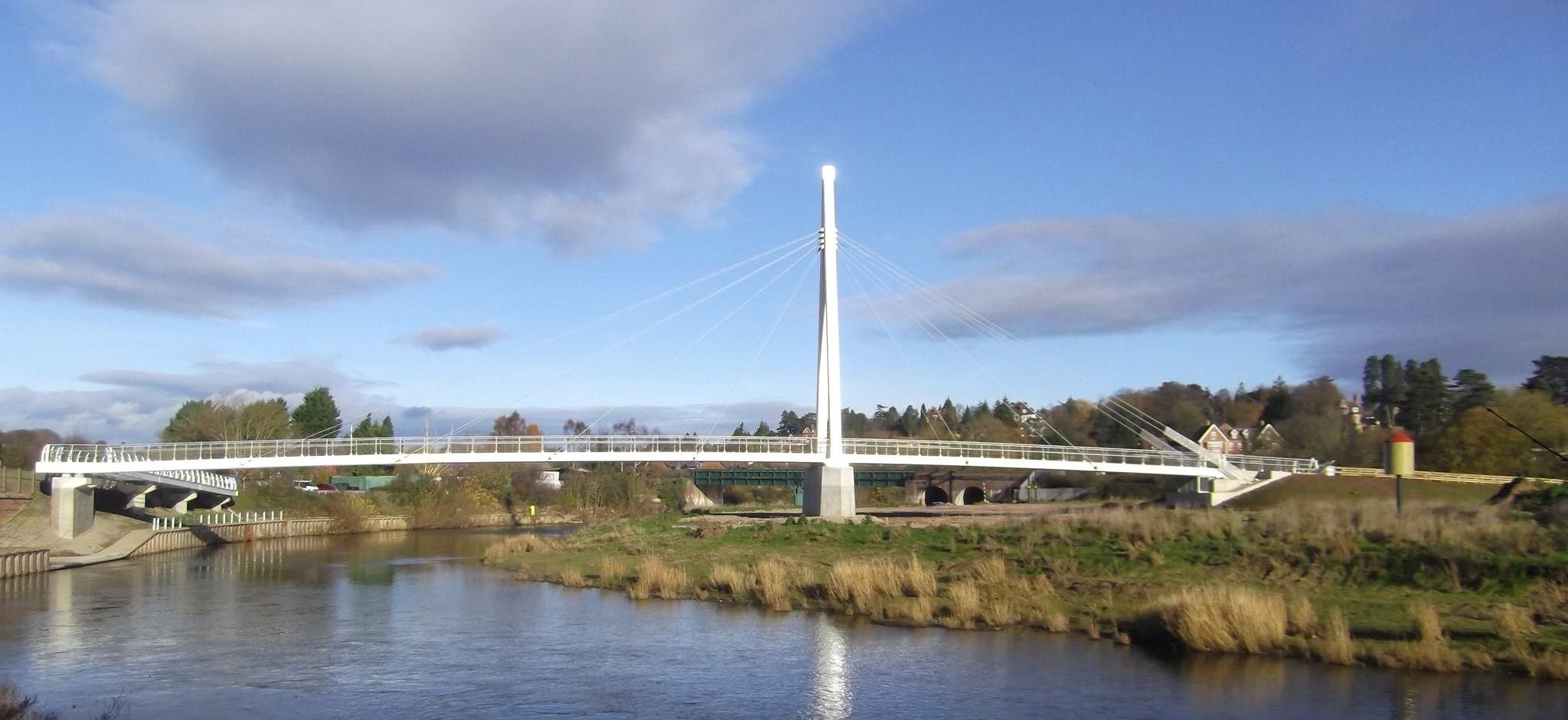A modern white suspension bridge over a river with trees, houses, and a partly cloudy sky in the background.