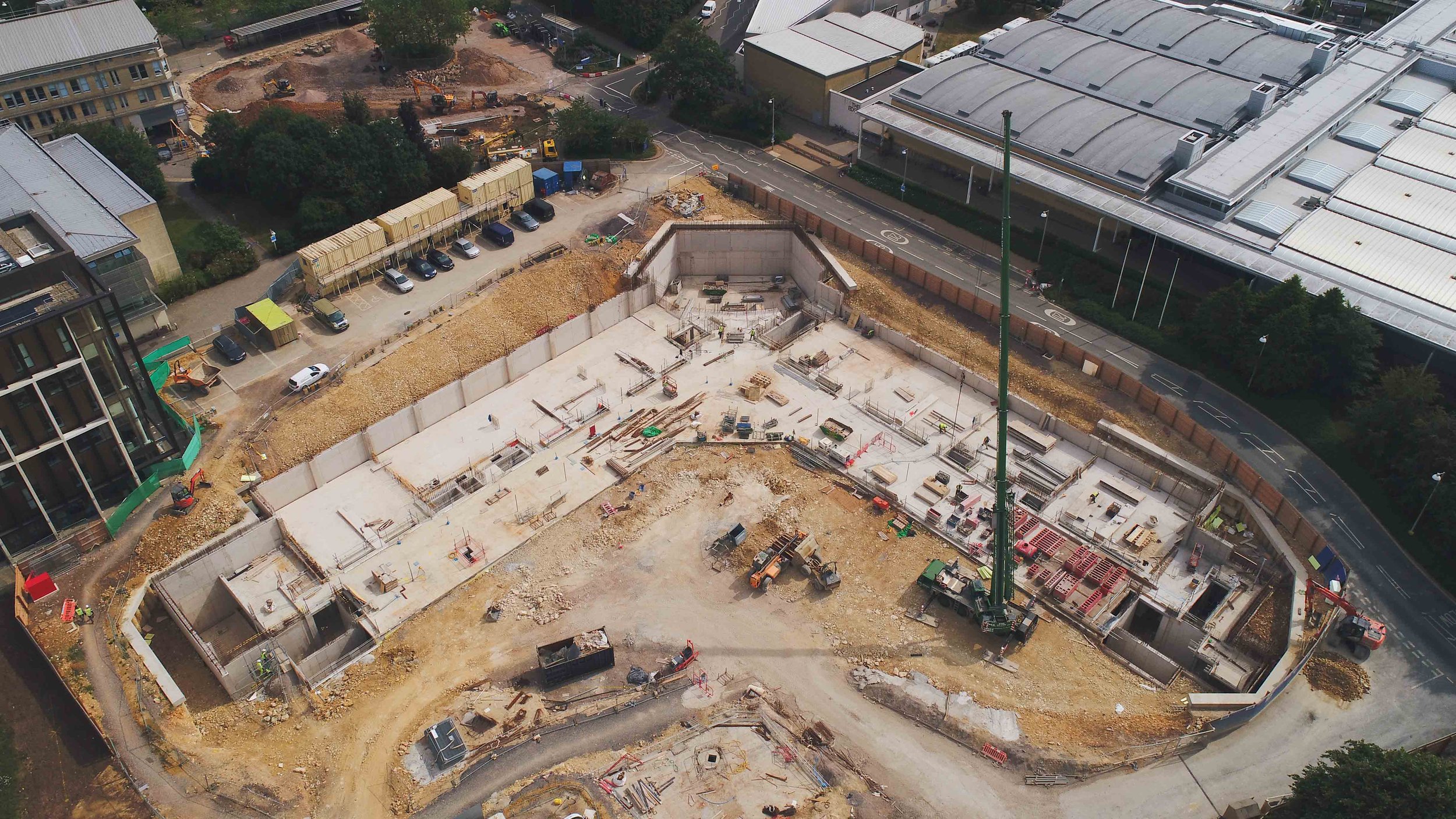 Aerial view of a construction site with partially built concrete structure, construction equipment, and materials, surrounded by office buildings and parking lot.