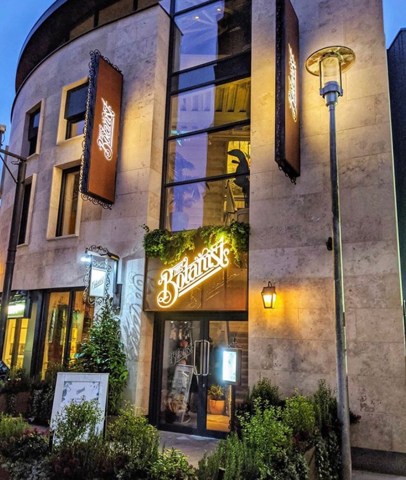 Exterior of "The Botanist" restaurant at night, featuring modern architecture, illuminated signage, and decorative greenery.