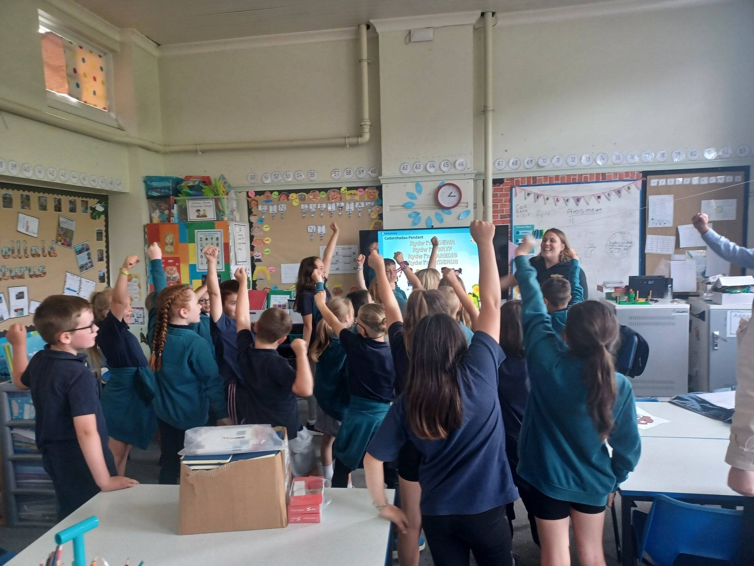 A classroom filled with young students raising their hands while a teacher smiles at the front of the room.