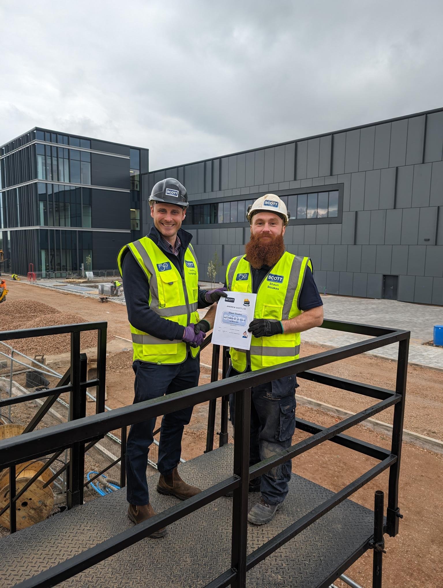 Two construction workers wearing yellow safety vests and helmets shaking hands on a construction site, holding a certificate, with a modern building in the background.