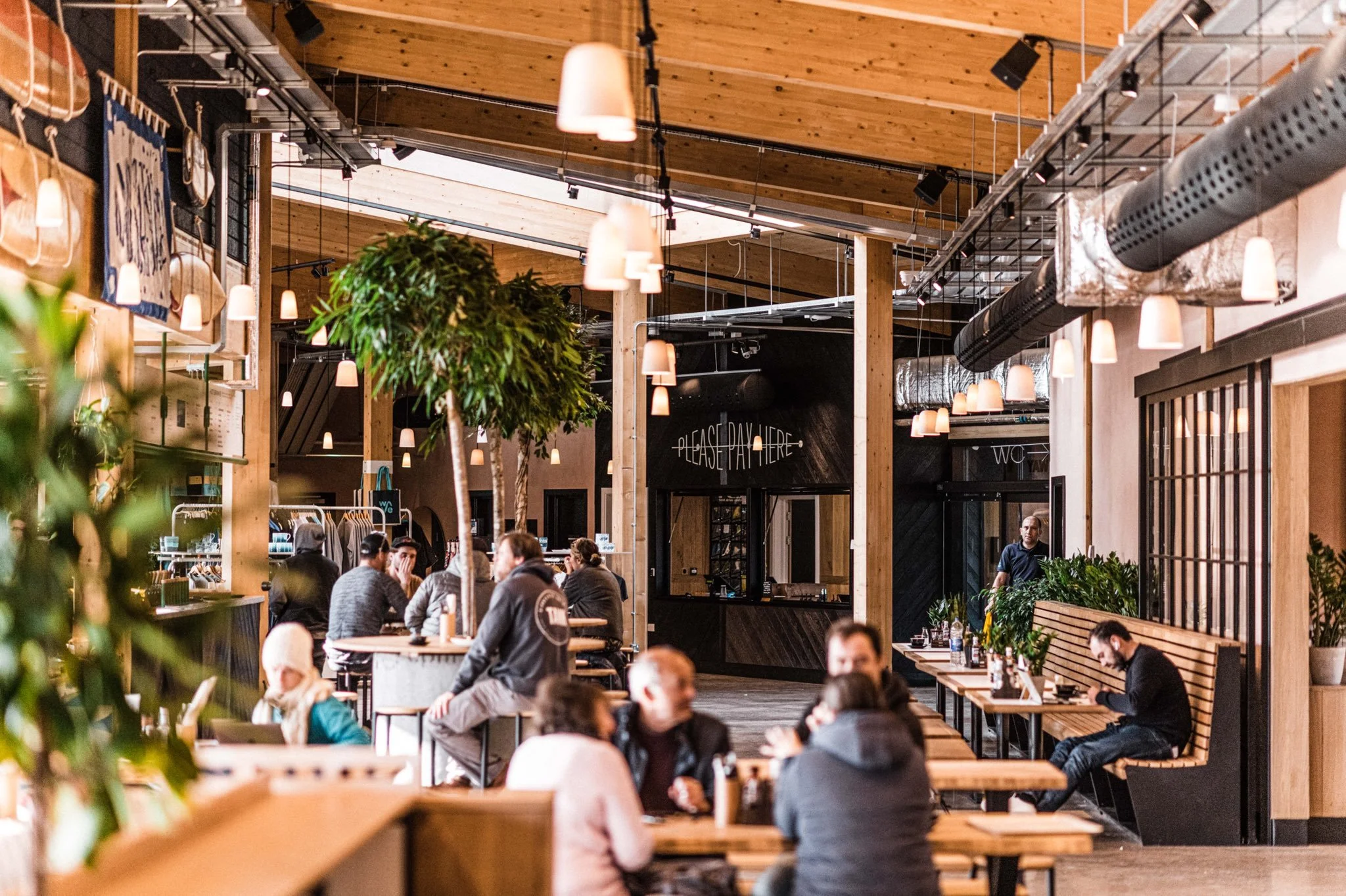 Interior of a modern café with wooden tables, hanging pendant lights, plants, and people sitting and eating.