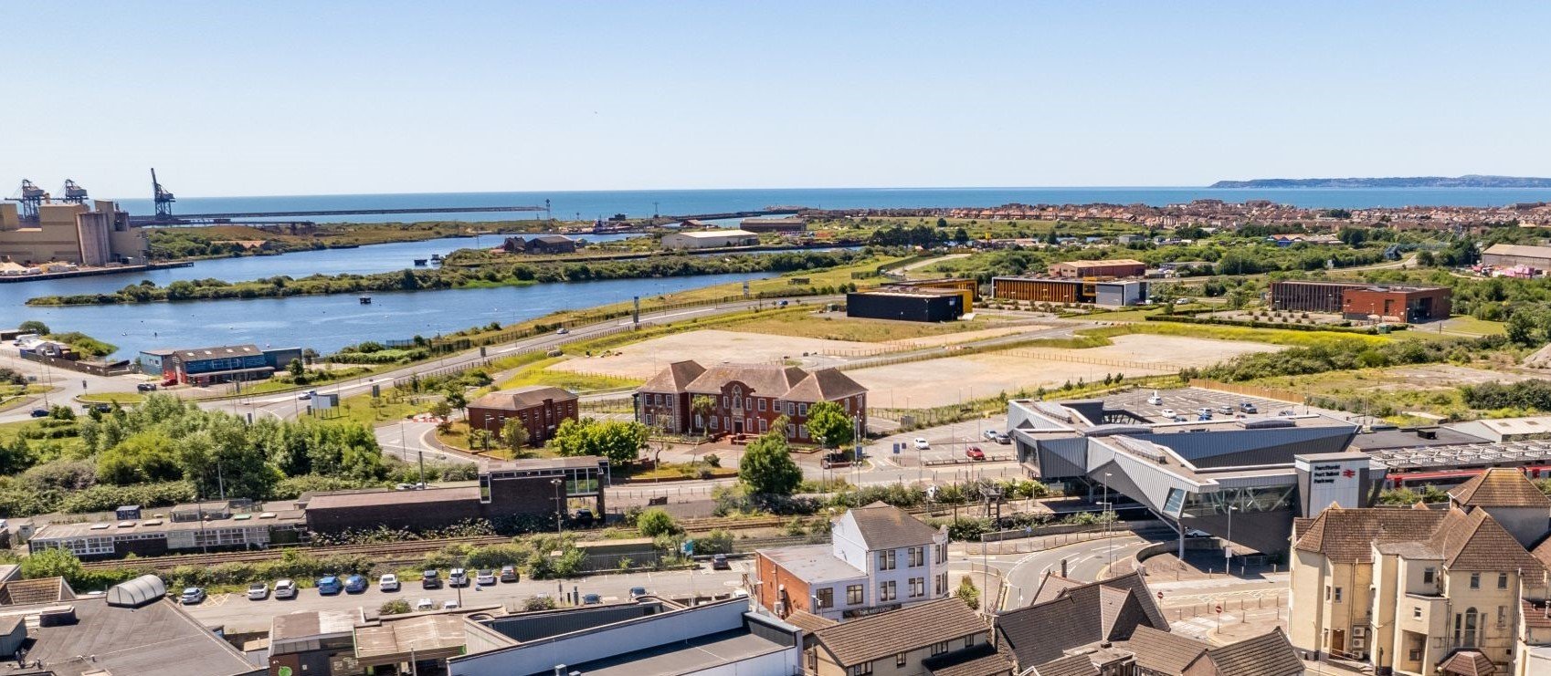 A cityscape featuring buildings, a river, green spaces, and a distant coastline with blue sky above.
