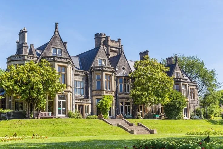 A large, historic stone mansion with multiple turrets and steeply pitched roofs, surrounded by a well-maintained lawn and trees, under a clear blue sky.