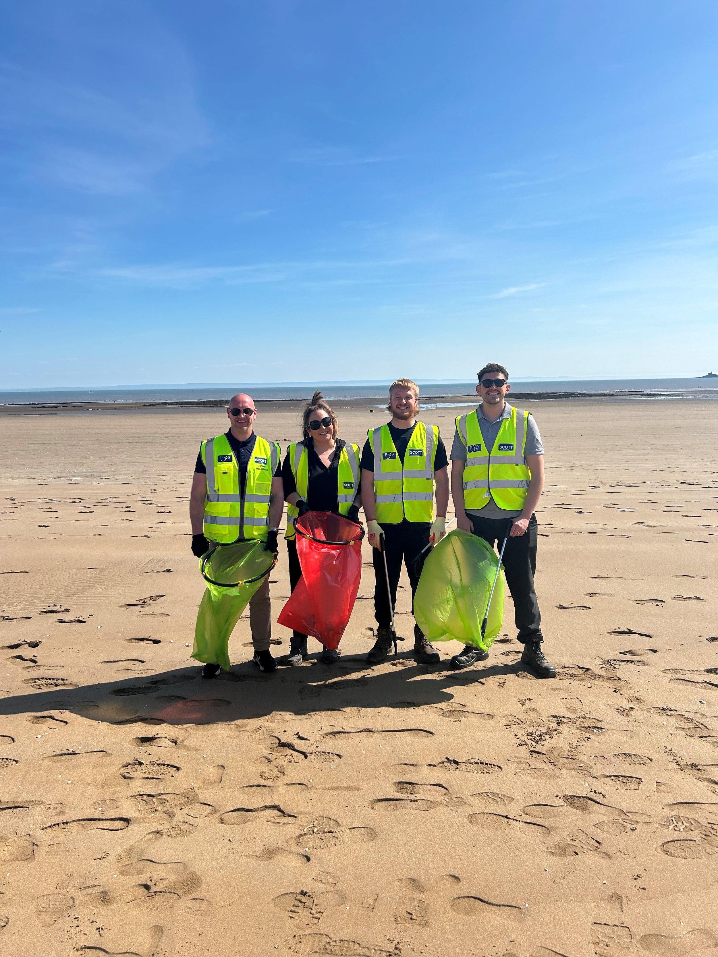 Four people in yellow safety vests trash collecting on a beach, holding trash bags, with footprints on the sand and the ocean in the background.