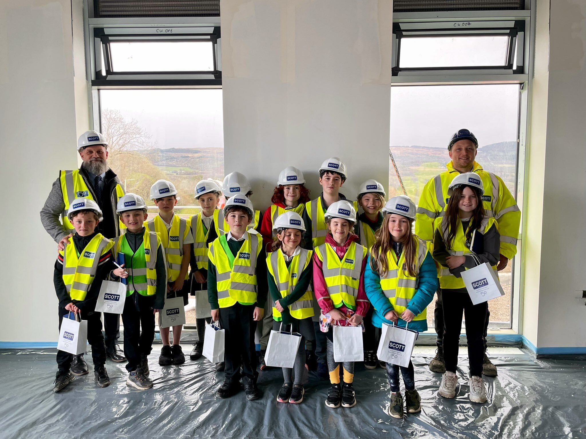 Group of children and two adults wearing yellow safety vests and white hard hats inside a building under construction, with large windows showing outdoor scenery.